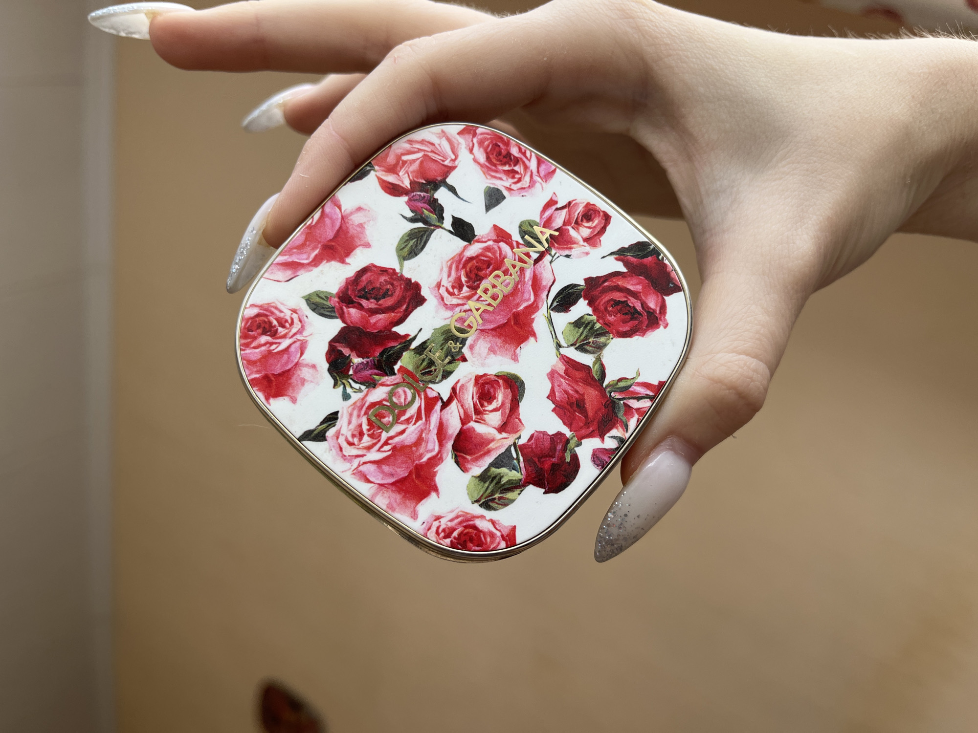 A person's hand with long, manicured nails adorned with glitter is holding a square compact with a floral pattern featuring red and pink roses. The compact has a gold-toned edge and embossed gold lettering that appears to read "DOLCE & GABBANA". The background is a plain, light brown color. There is no identifiable location in Ivanovo, Russia, or any other city.