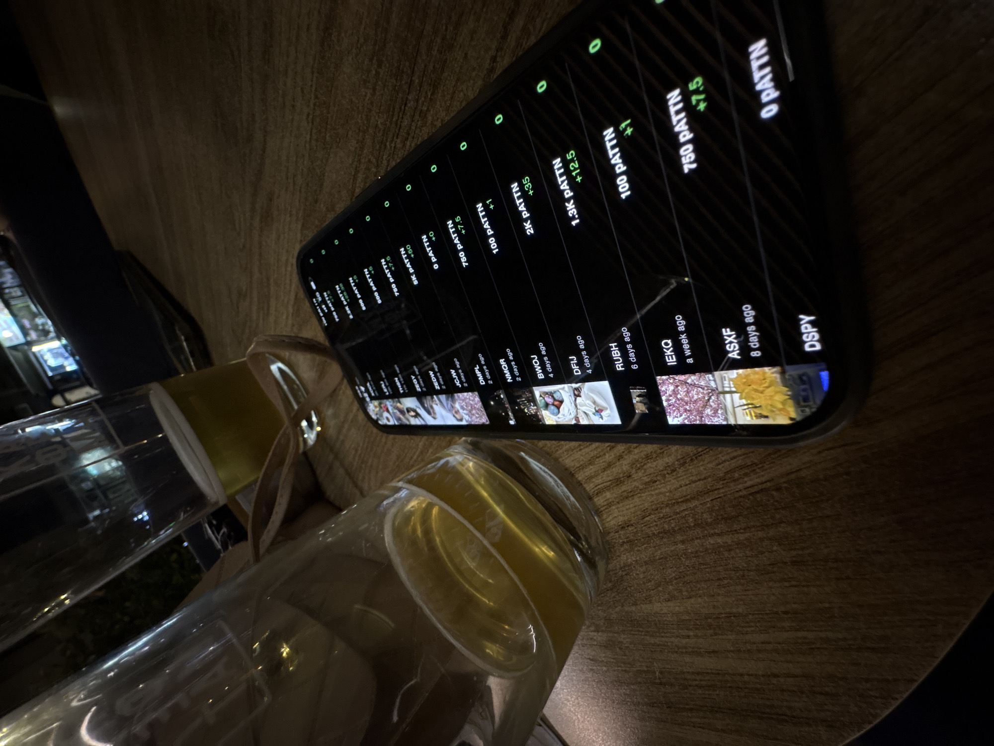 The image captures a close-up, angled view of a smartphone lying on a wooden table, with two glass bottles/jars in the foreground. One bottle contains a yellow liquid, while the other appears to hold a clear liquid or is mostly empty. The wooden table has a visible grain texture.
The smartphone screen is illuminated and displays a list-like interface with a dark background and green and white text. The content seems to be an application showing data, possibly related to finances, a game, or a tracking system. Entries include quantities labeled "PATTN" (e.g., "0 PATTN", "100 PATTN", "750 PATTN", "1.3K PATTN", "2K PATTN", "8K PATTN", "1.2K PATTN", "500 PATTN"). Many entries show numerical changes like "+7.5", "+1", "+12.5", "+35", "+50", and "+0". Timestamps or durations such as "2 days ago", "4 days ago", "6 days ago", "a week ago", and "8 days ago" are also visible. Various codes or names like "DSPY", "ASXF", "IEKQ", "RUBH", "DFIJ", "BWOJ", "NMQR", "DMPL", "JCJW", "ZEDW", "ZEGY", and "THON" are listed, often accompanied by small thumbnail images. Several "0"s are aligned at the top of the screen.
The scene is dimly lit, suggesting it is evening or night. The focus is on the objects on the table, likely in an indoor setting such as a restaurant or home in Kragujevac, Serbia.
