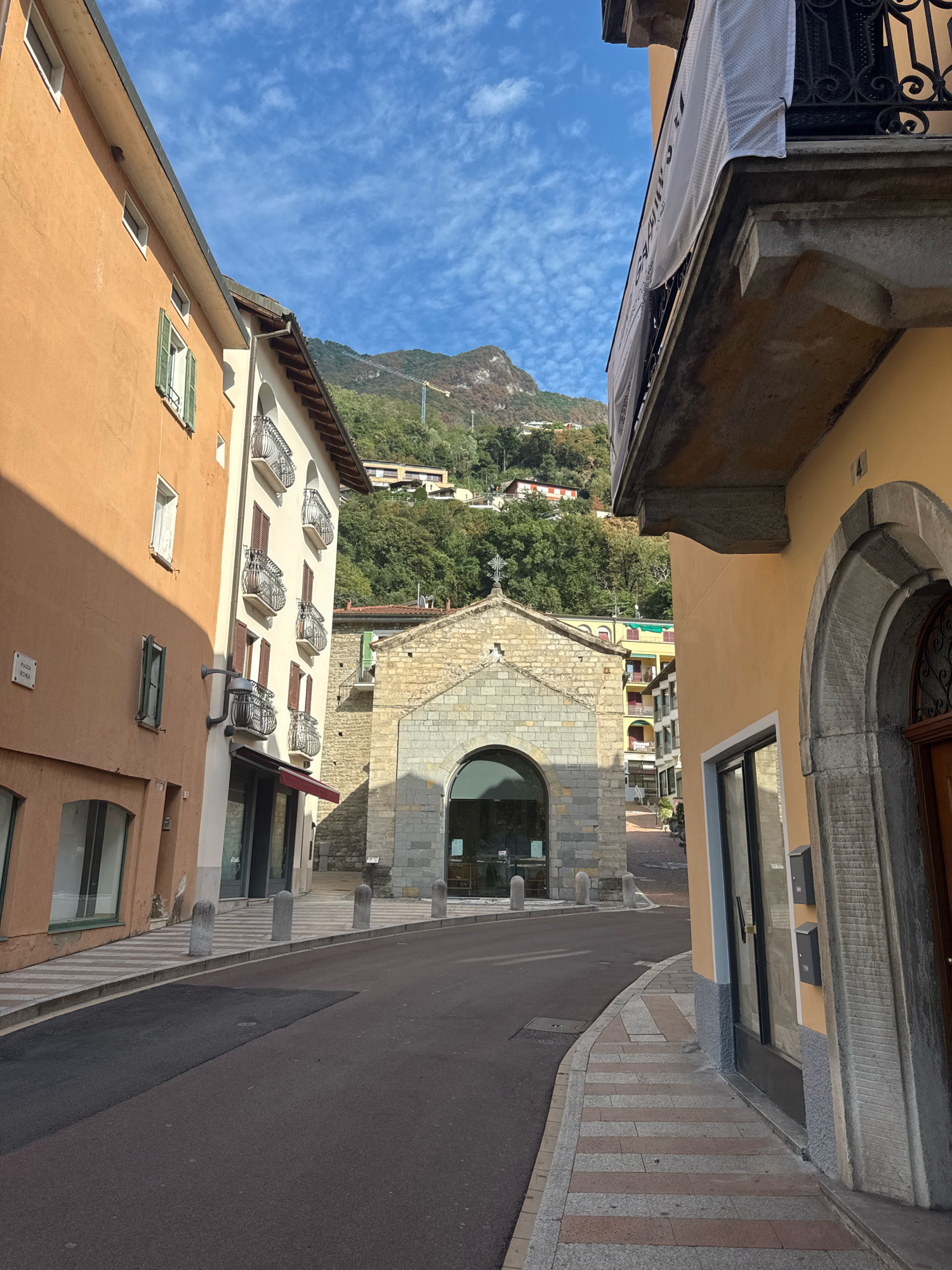A street scene in Campione d'Italia, Italy, features a narrow asphalt road curving towards a stone church with a cross on its roof. On the left, an orange building with several windows and green shutters stands tall, casting a shadow on the sidewalk. Next to it, a cream-colored building has multiple balconies with ornate railings. On the right, a yellow building with an arched entrance and decorative stonework is visible. The background reveals a lush green mountainside dotted with buildings and a crane. The sky above is a clear blue with scattered white clouds. The sidewalks are paved with a pattern of light and dark gray and reddish-brown stones. Several cylindrical stone bollards are placed along the sidewalk edge. A white banner with text is draped over a balcony on the upper right portion of the image. The overall atmosphere suggests a sunny day in a European town.