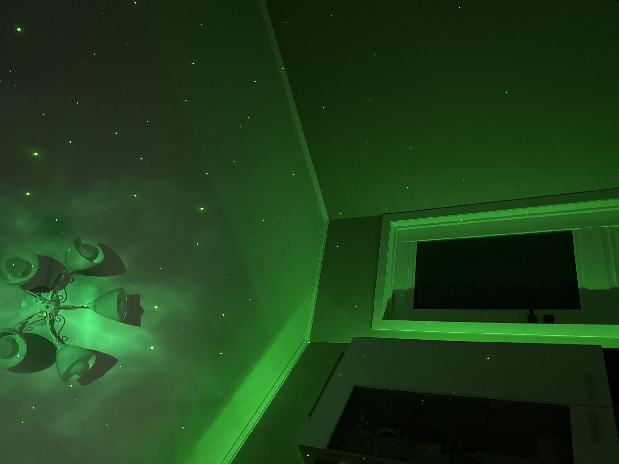 The image shows a corner of a room, illuminated with a bright green light that creates a starry effect on the walls and ceiling. A chandelier with multiple lamps is visible on the left side of the frame. The corner is formed by the ceiling and the wall, with white molding along the ceiling's edge. A rectangular window or display is positioned on the upper right side, showing a dark interior with a grid-like pattern. Below the window or display is a cabinet or shelf with a partially visible reflection on its surface. The overall lighting condition makes it difficult to discern specific details and colors. The location is Unknown, Unknown.