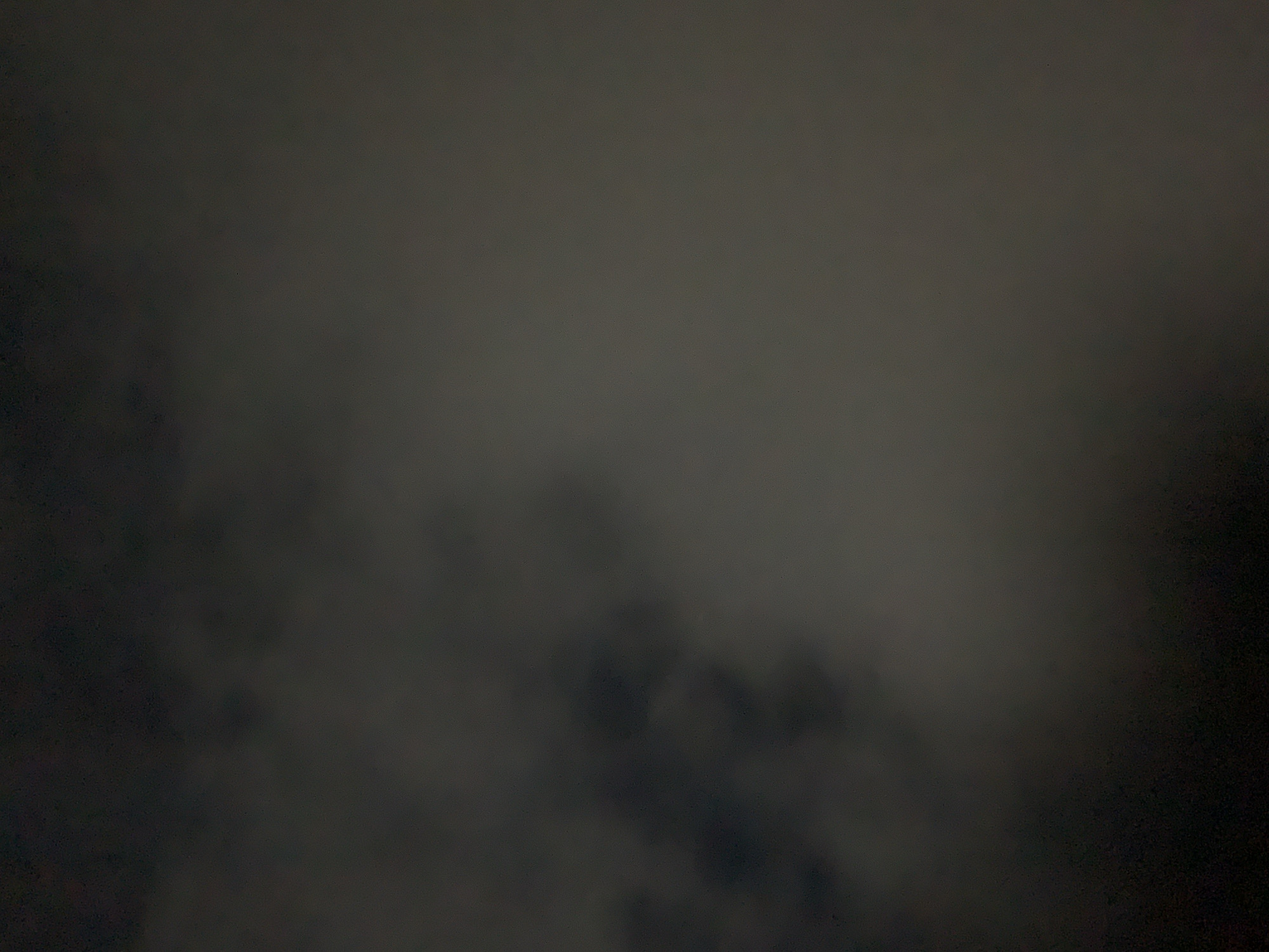 The image captures a dark, heavily overcast night sky above New Stadium, Nigeria. Dense, dark gray to black cloud formations occupy the left portion of the frame, showing distinct, billowy structures. Toward the right, the cloud cover is slightly lighter and more diffuse, presenting a softer gradient of dark gray. The scene is characterized by extremely low illumination, with no discernible light sources, celestial bodies, or terrestrial features. The texture of the sky suggests a thick, unbroken layer of clouds under deep night conditions. No individuals, objects, or specific actions are present in the frame, which is solely an atmospheric study.