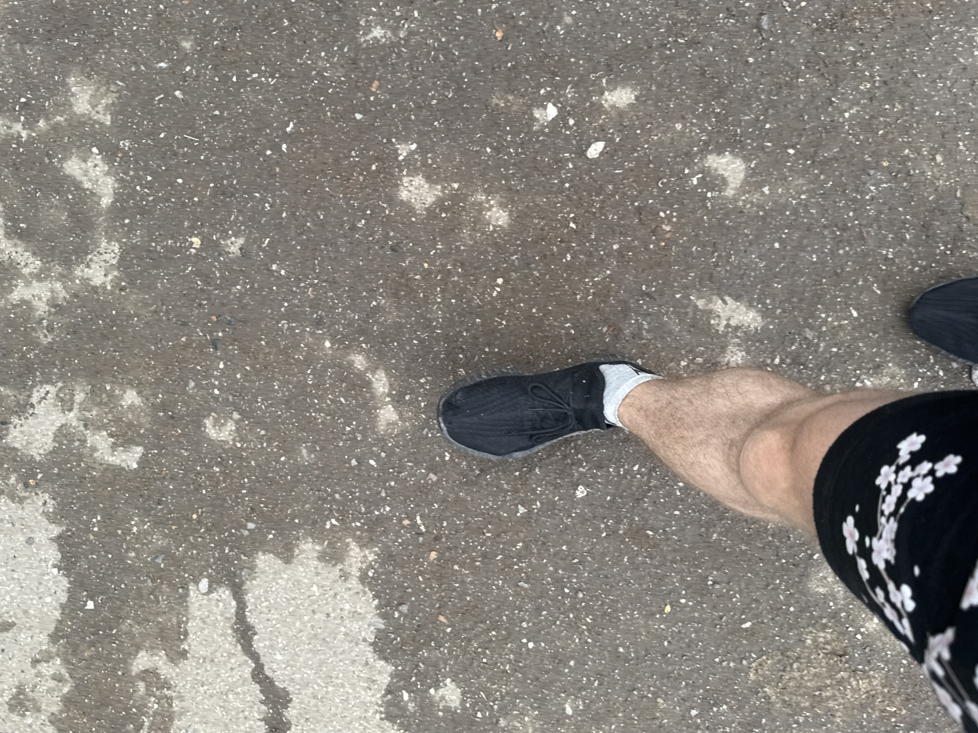 The image shows a first-person perspective of someone walking on a wet asphalt surface. The asphalt has patches of lighter colored material, possibly concrete or lighter asphalt, and is speckled with small stones and debris. The person is wearing a black athletic shoe with white laces and a white sock. A portion of their bare leg is visible above the sock, and they are wearing black shorts with a pattern of light pink cherry blossoms. The scene appears to be outdoors on a street or pathway, indicated by the urban ground texture. There is no information in the image to suggest it is in Oral, Kazakhstan.