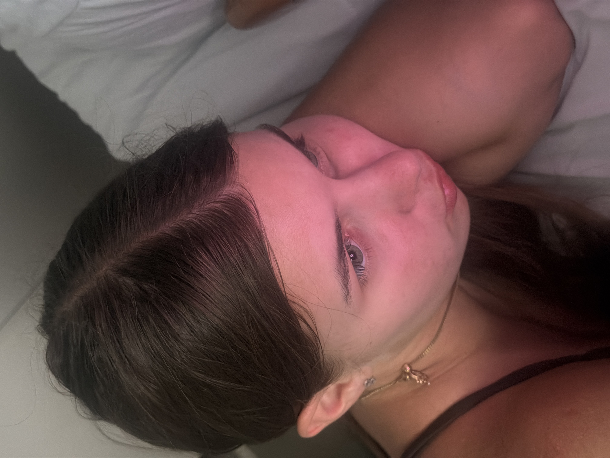 The image is an extreme close-up of a young Caucasian woman lying down, her head resting on a white surface, likely a pillow or bedding. Her face is angled away from the camera, with her left eye visible. She has dark brown hair, which appears to be long and slightly wavy. Her skin is illuminated with a pinkish hue, possibly from artificial lighting. She is wearing a dark-colored strap, suggesting she is wearing a top or dress. A thin gold necklace with a small pendant is visible around her neck. The background is a plain, muted color. The image does not contain any elements that suggest it is located in Nha Trang, Vietnam, or any other specific city.