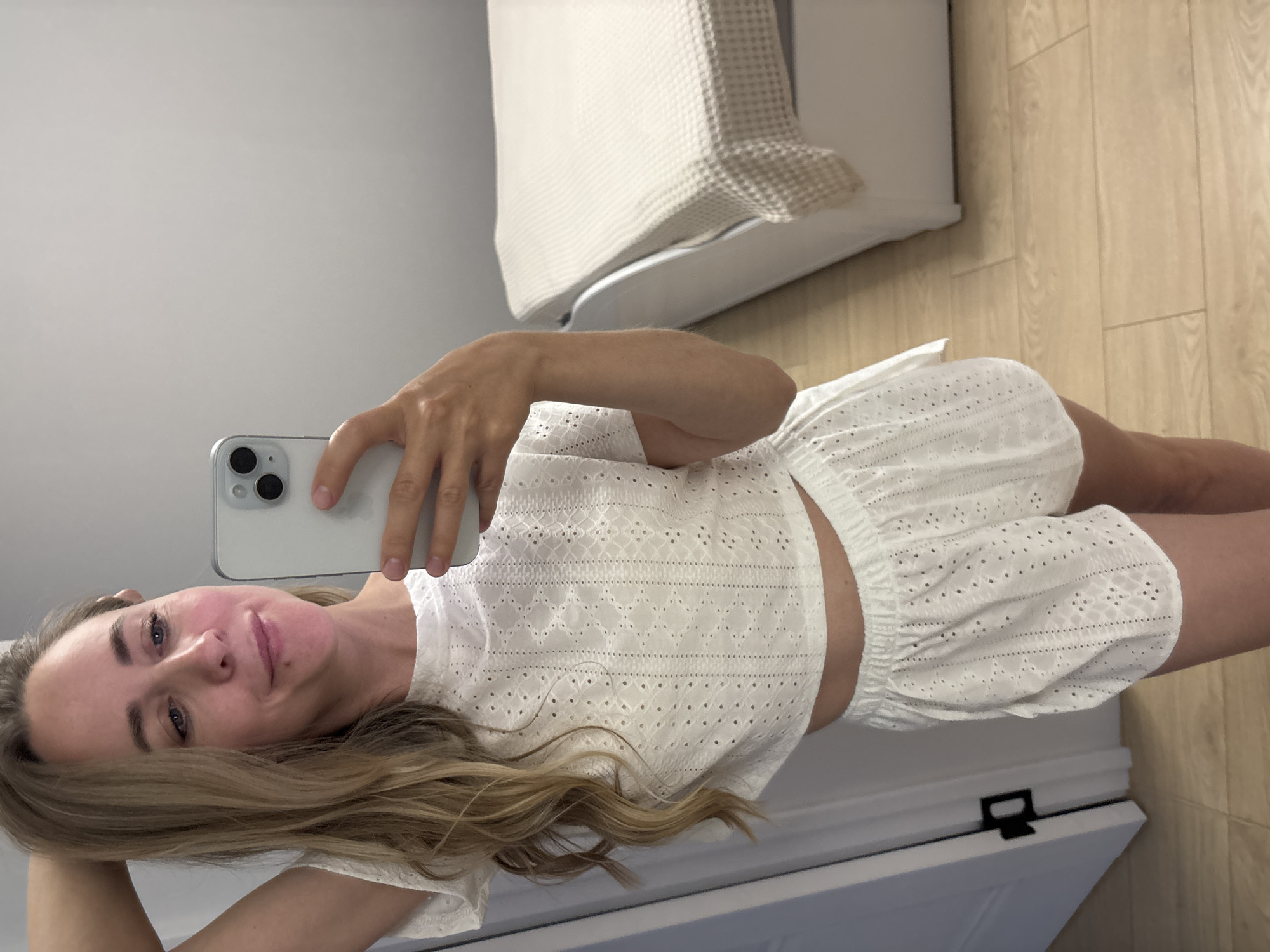 A young woman is taking a selfie indoors. She is wearing a white eyelet-style crop top and matching shorts. She holds a white smartphone in her right hand. The background includes a bed with white bedding, light wood flooring, and a white door. The location is not specified to be Chaikovsky, Russia.