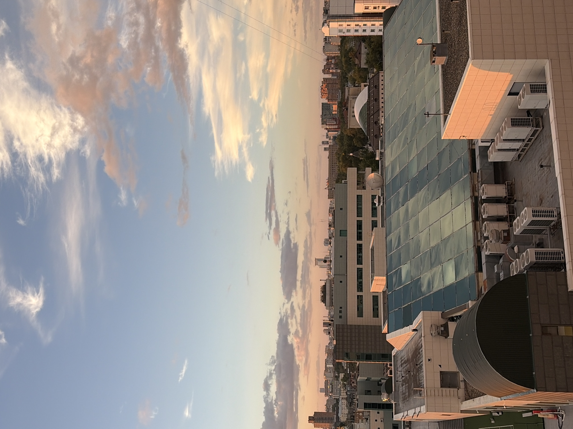 The image depicts a panoramic view of a cityscape, likely Moscow, Russia, during the golden hour. The sky is a blend of pale blue and soft pinks, with wispy clouds illuminated by the setting sun. In the foreground, a rooftop scene is visible, featuring various architectural elements. These include a large, multi-paneled glass roof, possibly part of a stadium or large building, reflecting the sky. Adjacent to it is a building with a beige, tiled facade, partially bathed in warm sunlight. Several air conditioning units are neatly arranged on the rooftops, indicating an urban environment with modern infrastructure. In the mid-ground and background, a dense collection of buildings, ranging from mid-rise structures to taller skyscrapers, stretches across the horizon, interspersed with trees. The overall impression is one of a bustling city at dusk, with a blend of commercial and residential architecture under a tranquil sky. There are no people visible in the image.