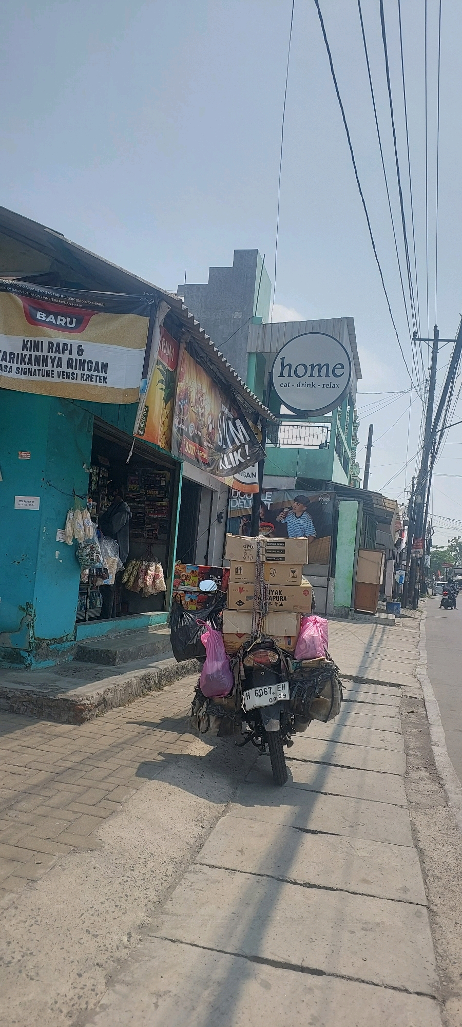 The image captures a street scene in Semarang, Indonesia, on a bright, sunny day. In the foreground, a motorcycle is heavily loaded with various goods, including several cardboard boxes stacked high and two large pink plastic bags. The motorcycle has license plate number H 6067 EH. The motorcycle is parked on a sidewalk constructed of paving stones and concrete. Adjacent to the sidewalk is a street.
To the left of the sidewalk is a row of buildings. The building closest to the motorcycle appears to be a small shop with a turquoise-colored facade. A banner with Indonesian text, partially obscured, is hung above the shop entrance. Another shop to the right has a circular sign with the word "home" and the tagline "eat - drink - relax" in English. The buildings have a slightly worn appearance, consistent with an urban environment.
In the background, electrical wires crisscross the sky, and other buildings and a utility pole are visible. A few other motorcycles and possibly cars are seen further down the street, suggesting ongoing activity. The overall impression is of a typical commercial street in a bustling Indonesian city.