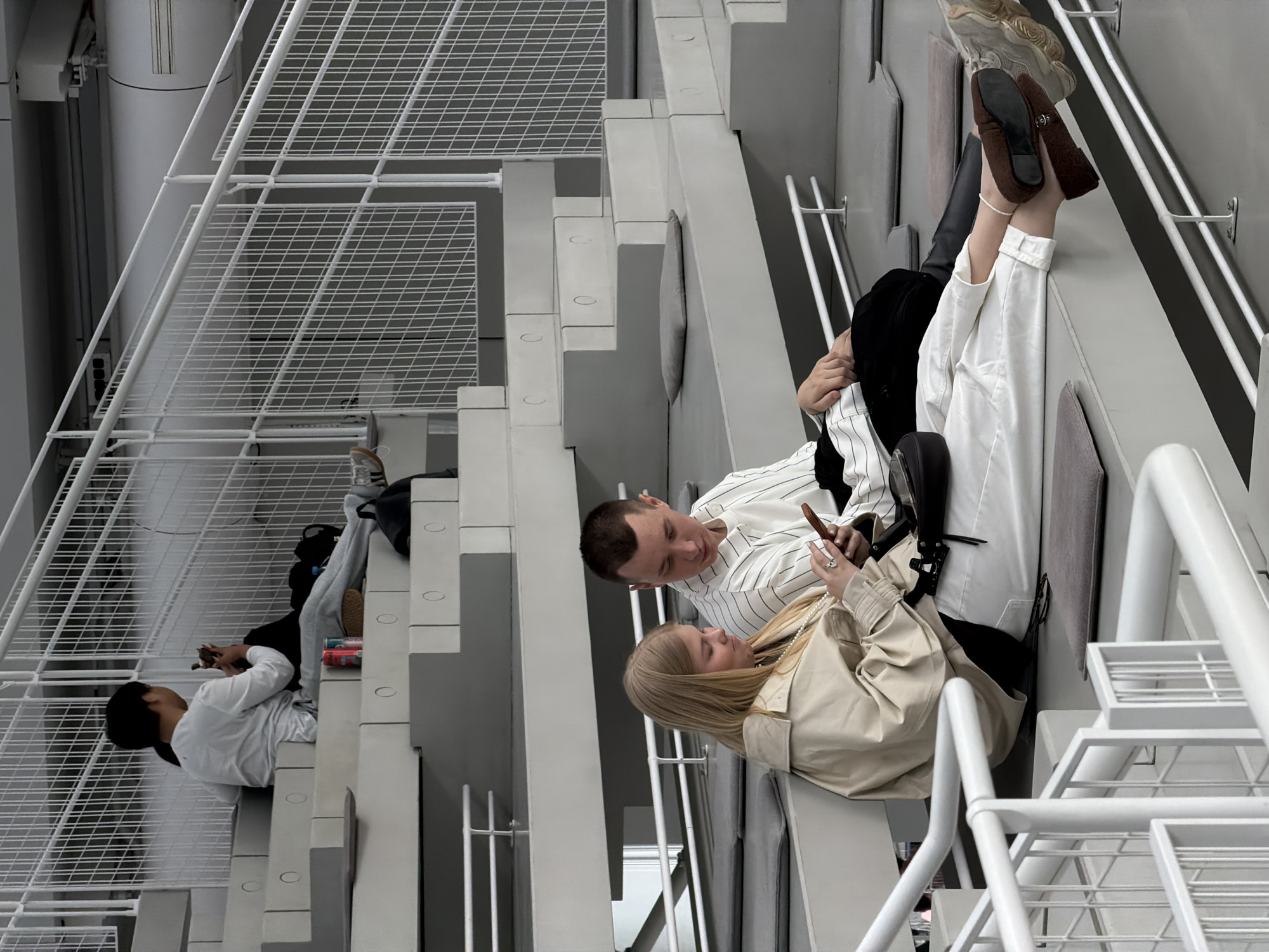 Three individuals occupy the staircase of the Garage Museum of Contemporary Art in Moscow, Russia. A couple, situated halfway up the stairs, is focused on a mobile phone. The woman, wearing a tan jacket, sits next to a man wearing a striped shirt. A second man, wearing a white shirt, sits on the lower stairs. The staircase is white, with gray pads on each step, and it features a white metal mesh design.