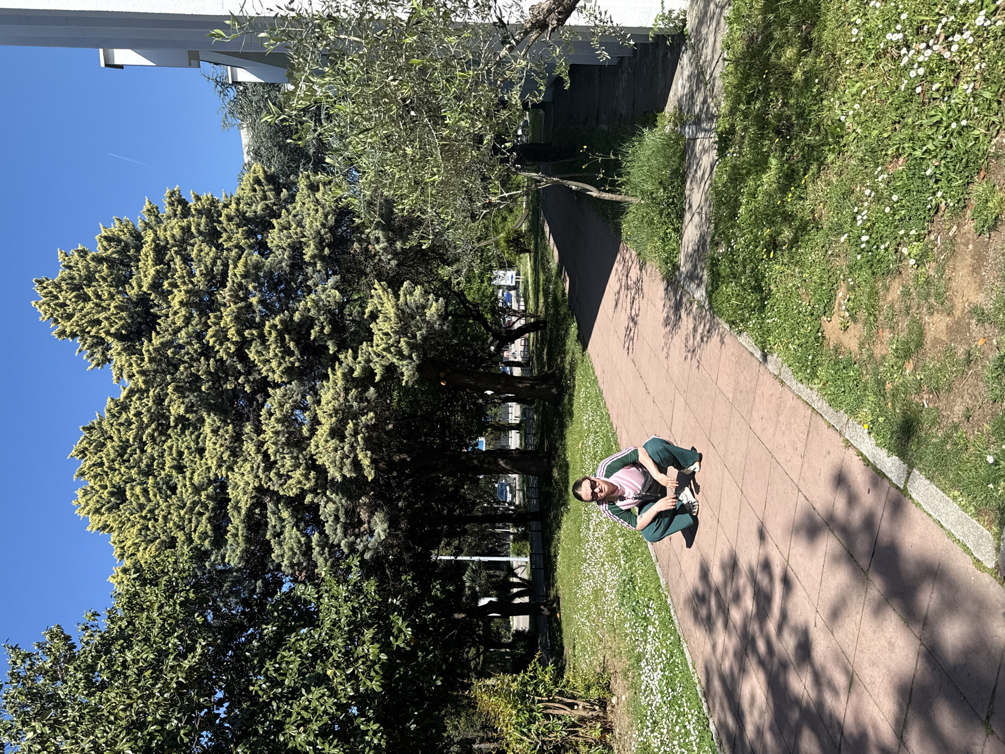 A person is squatting on a brick walkway in a park-like setting in Bar, Montenegro. The walkway is surrounded by grass and trees, and there are buildings visible in the background. The sky is clear and blue. The person is wearing a pink and green athletic suit and sunglasses. There are dandelions and other small white flowers growing beside the walkway. The image is taken at an angle, giving a wide view of the scene.