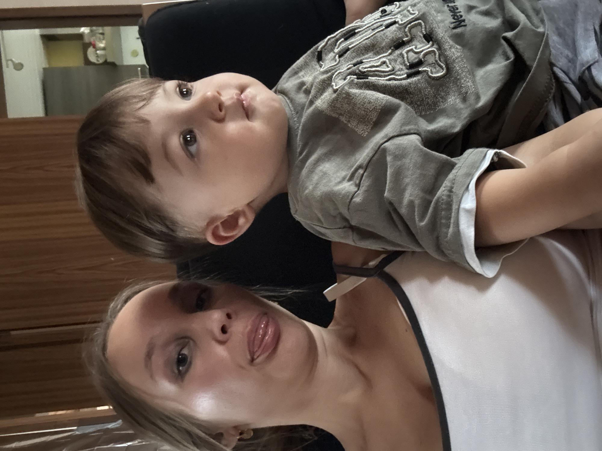 The image shows two people, a woman and a young child, indoors. The woman is on the left, with her face turned towards the camera and her tongue sticking out. She is wearing a white tank top. The child is on the right, facing away from the camera and looking to the right. The child is wearing a grey t-shirt. The background appears to be a room with wooden paneling on the walls. The scene is dimly lit. There is no information in the image to suggest the location is in Überherrn, Germany.