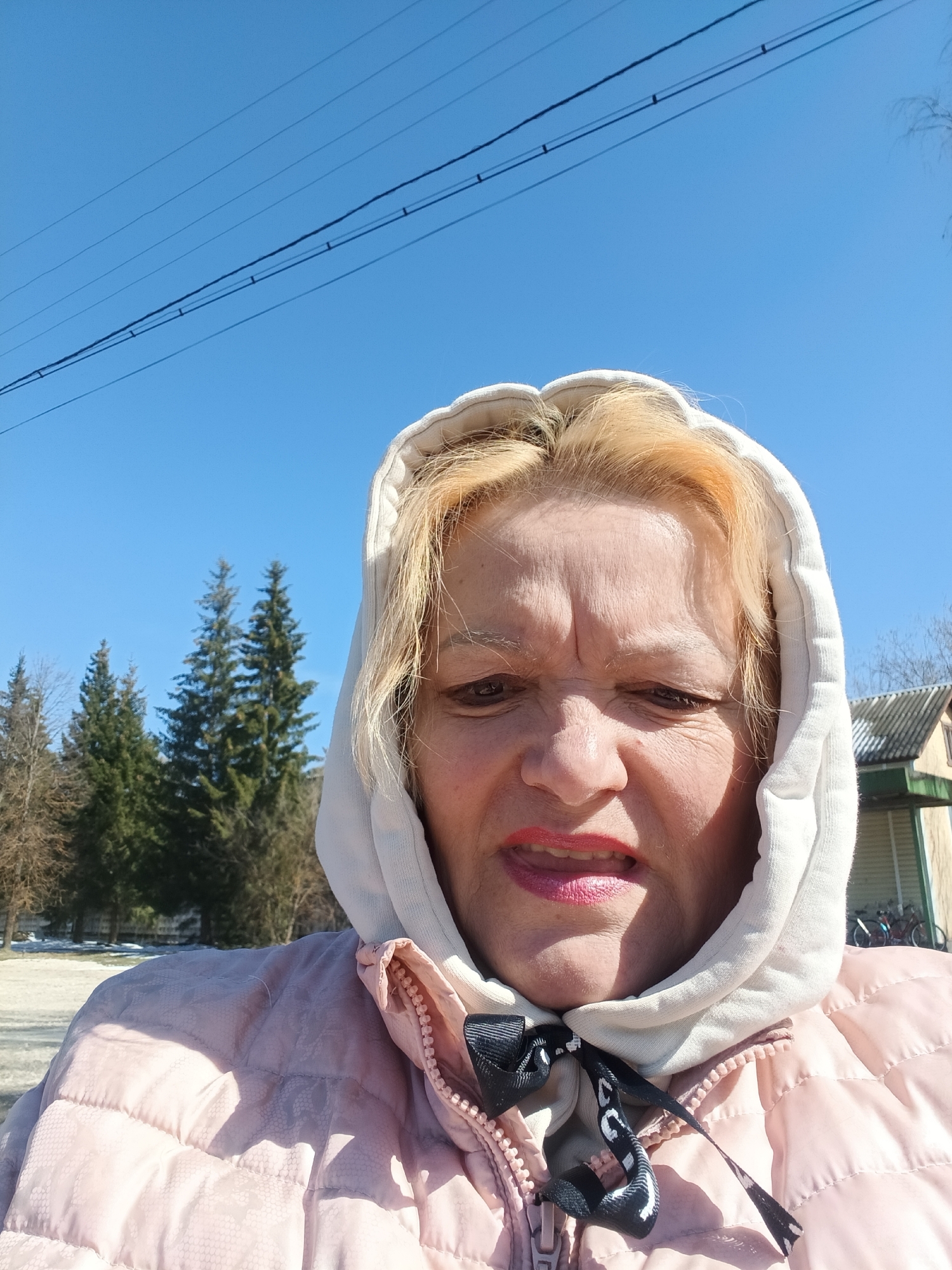 This media file features a close-up, slightly upward-angled portrait of a woman taken outdoors on a clear, sunny day. The subject is a woman appearing to be middle-aged or older, with blonde hair, looking directly at the camera with a squinting or grimacing expression. She is wearing a light-colored (possibly off-white) hooded sweatshirt with the hood up, layered under a light pink quilted puffer jacket. A black and white patterned ribbon or tie is visible around her neck, fastened over the jacket's zipper. Her lips are painted with bright red lipstick and are slightly parted.
The setting is an outdoor environment, possibly rural or semi-rural, given the natural elements and structures. In the background to the left, several tall coniferous trees stand alongside some bare deciduous trees. The ground shows patches of snow or ice, indicating that the photo was taken in late winter or early spring. To the right, part of a building with a grey corrugated roof and light-colored walls is visible in the distance. Above the woman's head, utility lines (power lines) stretch across a vibrant, clear blue sky. The strong sunlight suggests it is midday. No discernible text is visible within the image, though the ribbon has an illegible pattern. The context for the location is Красный Маяк, Russia.