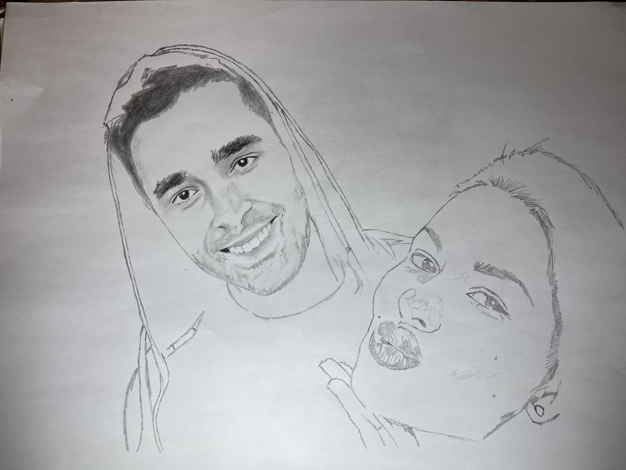 The image displays a monochrome pencil drawing rendered on a white sheet of paper, oriented diagonally. The drawing features two human faces. The prominent upper face, positioned to the right of the canvas, is that of a male with short, dark hair, a trimmed beard, and a clear, smiling expression, directly engaging the viewer. He appears to wear a light-toned hooded garment or cloth draped over his head and shoulders. Below and partially to the left of the male's face is a second, smaller face, likely a younger individual or child, characterized by lighter, possibly styled, hair and an upward gaze with slightly parted lips. A faint, partial outline of a hand is visible near the lower subject's left cheek. The artwork employs pencil shading to delineate features, hair, and clothing folds. The depicted scene is an artistic portrait and contains no specific geographical landmarks, architectural elements, or environmental cues that would indicate a physical location within the city of Jos, Nigeria.