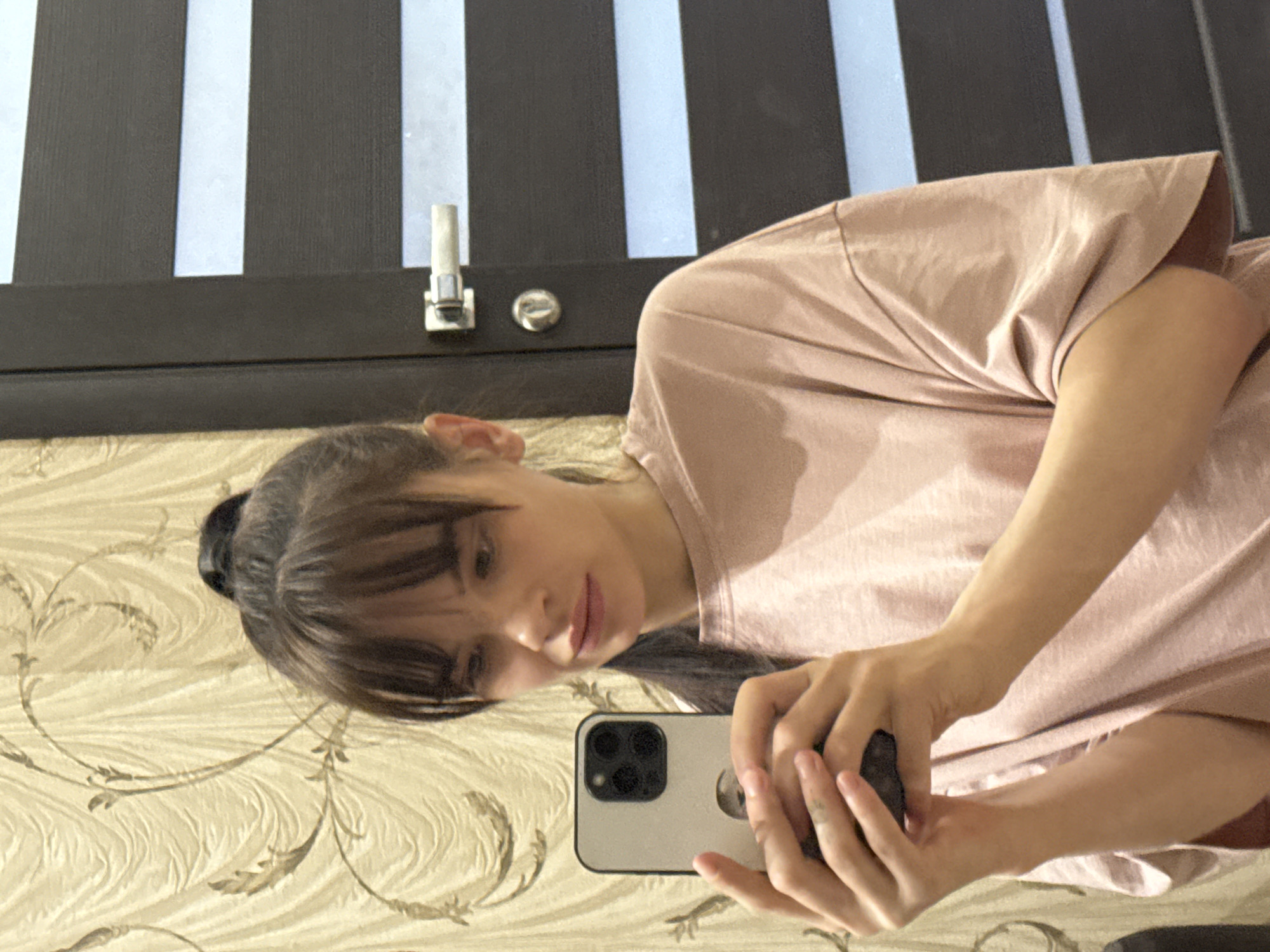 A woman is taking a selfie with her iPhone in a room. The room has wallpaper with a floral pattern and a dark brown door with white stripes. The location is unknown, there is no way to determine that it is Rostov-on-Don, Russia.