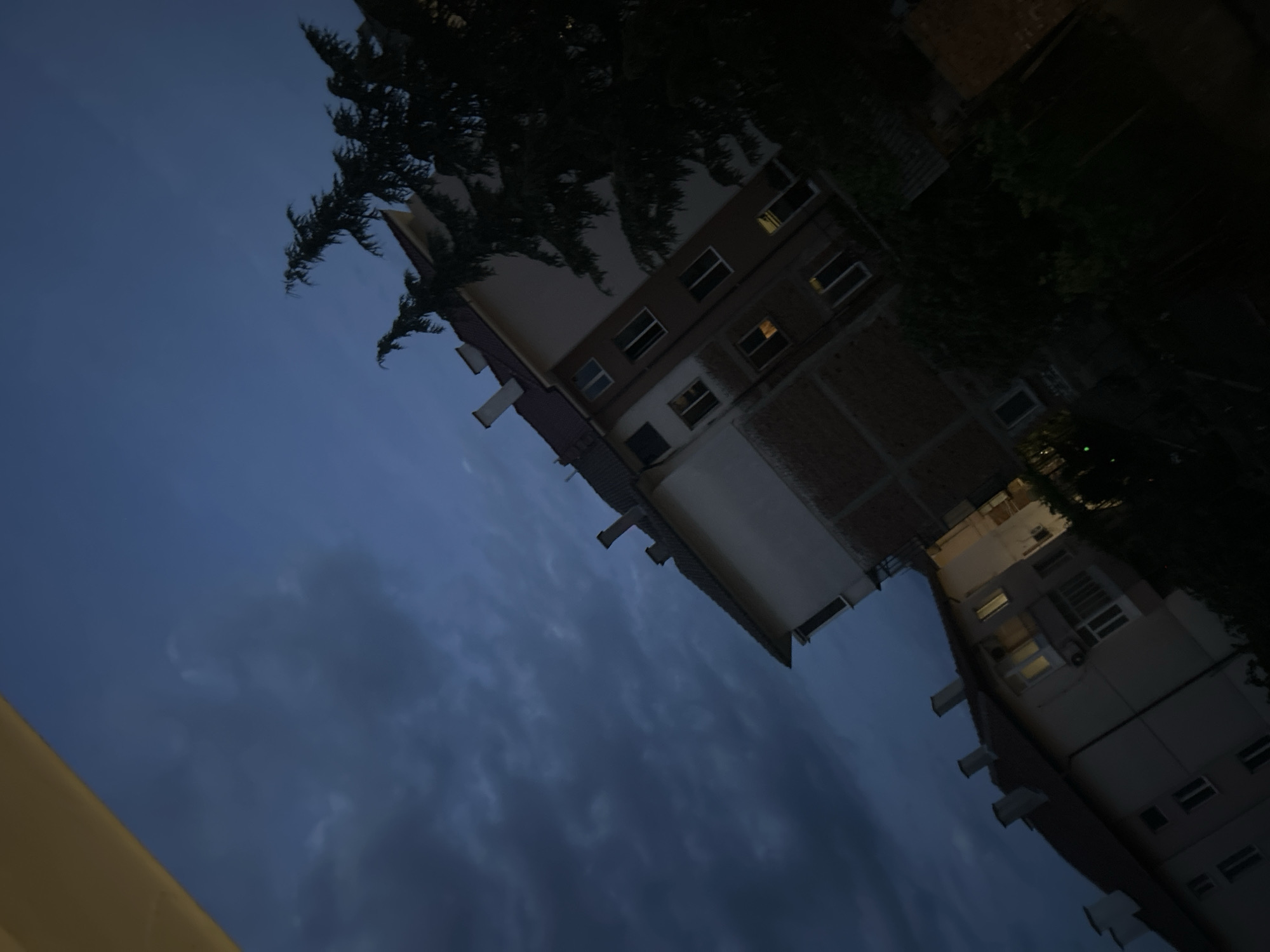 This is a photograph taken at twilight, with the sky a deep blue, filled with dark, textured clouds. In the foreground, angled from the bottom left to the top right, are the upper floors of apartment buildings. The buildings are light-colored with dark roofs and visible chimneys. Some windows are lit, casting a warm glow into the dim evening. A large, dark evergreen tree is visible on the right side of the image, partially obscuring the buildings and the sky. The overall impression is one of a quiet, urban evening with an approaching storm or the end of daylight.
