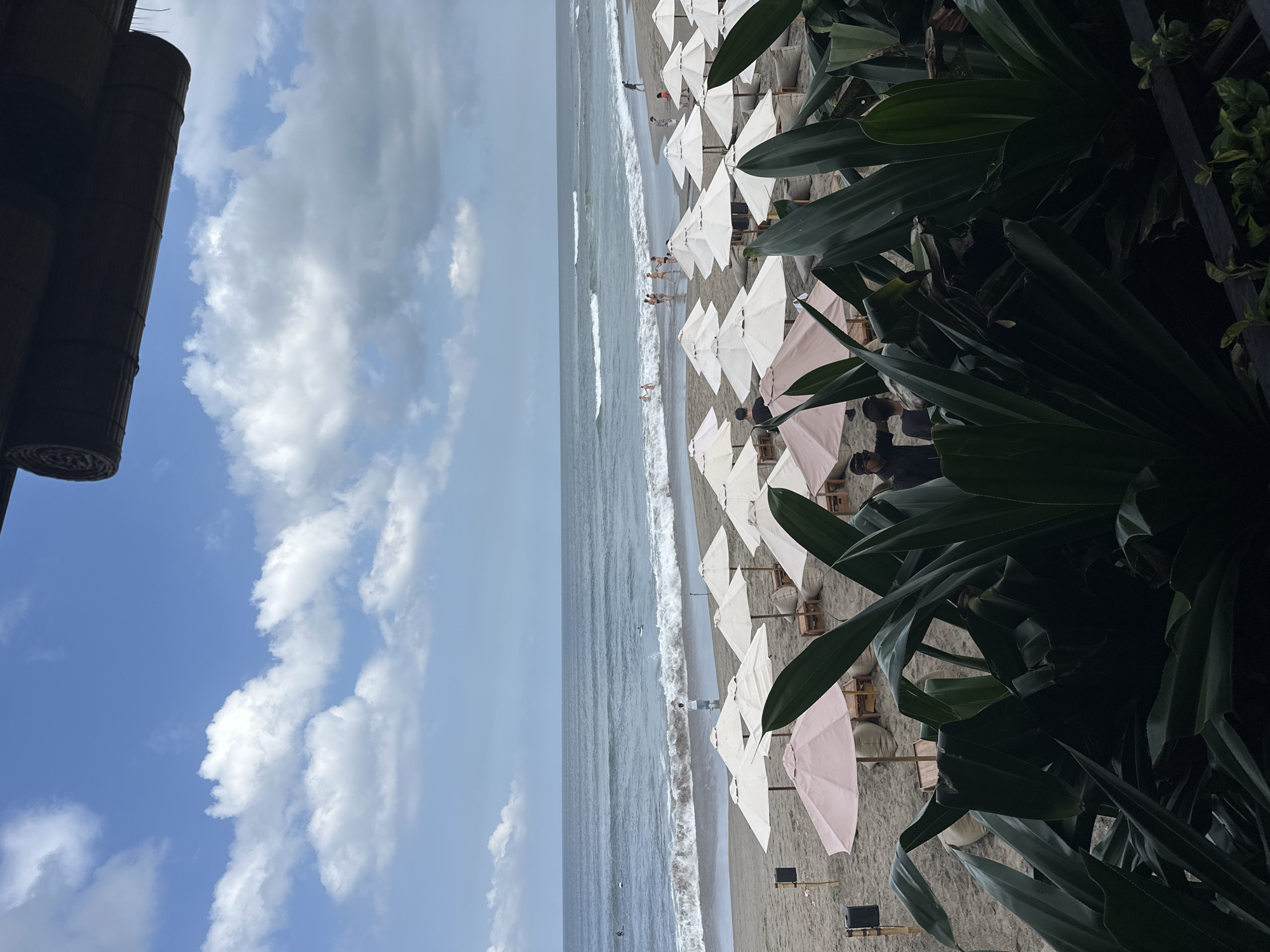 The image depicts a coastal scene at a beach in North Kuta, Indonesia. The beach is populated with numerous white and pale pink sun umbrellas arranged in rows along the sand. In the distance, people can be seen swimming or wading in the ocean. The water is visible with white waves. The sky is blue with scattered cumulus clouds. Vegetation, including broad, dark green leaves, appears in the right portion of the frame, and part of a structure that seems to be the roof of a building is on the top-left corner of the frame.