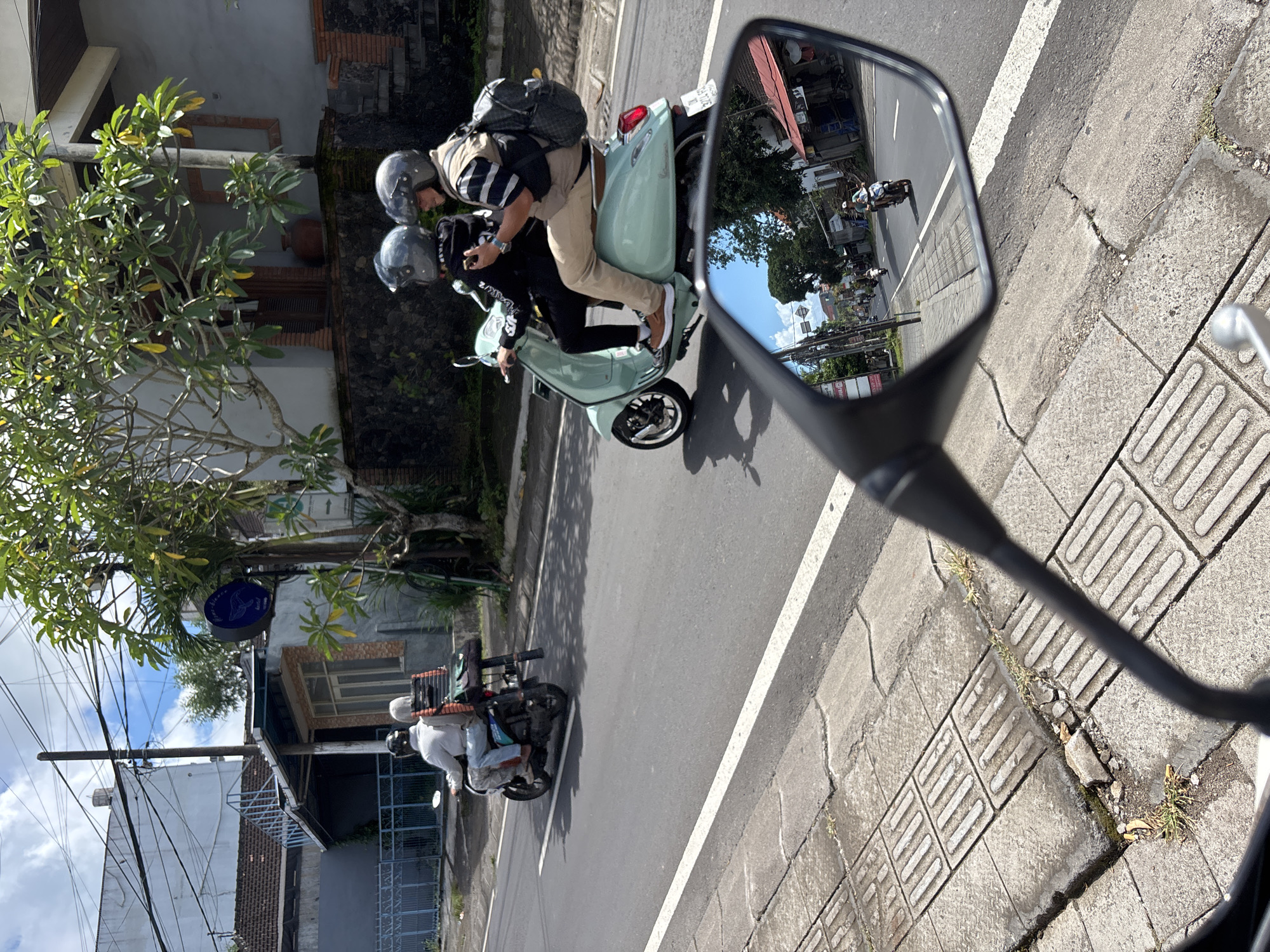 The scene is a street in Ungasan, Indonesia. The image is taken from the perspective of a motorcycle rider, with the motorcycle's side mirror visible in the foreground. Reflected in the mirror is a view of the road behind, showing another motorcycle carrying two helmeted individuals. The riders on the second motorcycle are both wearing helmets and casual clothing. One rider is wearing a light-colored shirt, while the second is wearing a black hoodie with designs. The motorcycle they are riding is a light-green Vespa-style scooter, with a large backpack strapped to the back. On the road, another motorcycle is also visible. The background includes buildings and trees typical of a tropical urban environment. The road appears to be well-maintained with a white line marking the lane. The sidewalk shows a textured surface with cracked and missing pieces. Overall, the scene captures a typical day of motorcycle traffic in Ungasan, Indonesia.