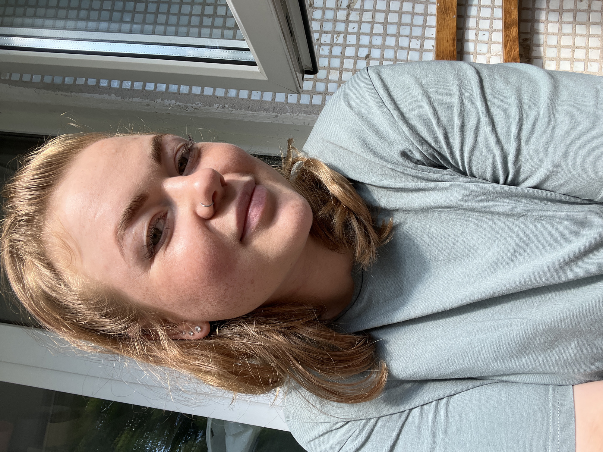 A medium shot shows a young woman with light brown hair, freckles, and a nose ring smiling slightly. She is wearing a light blue-green t-shirt. Her left ear has two small stud earrings. The background consists of a white framed window with a view of greenery and a tiled wall. The image appears to be taken outdoors on a sunny day. No specific location in Chaikovsky, Russia, is identifiable from the image.