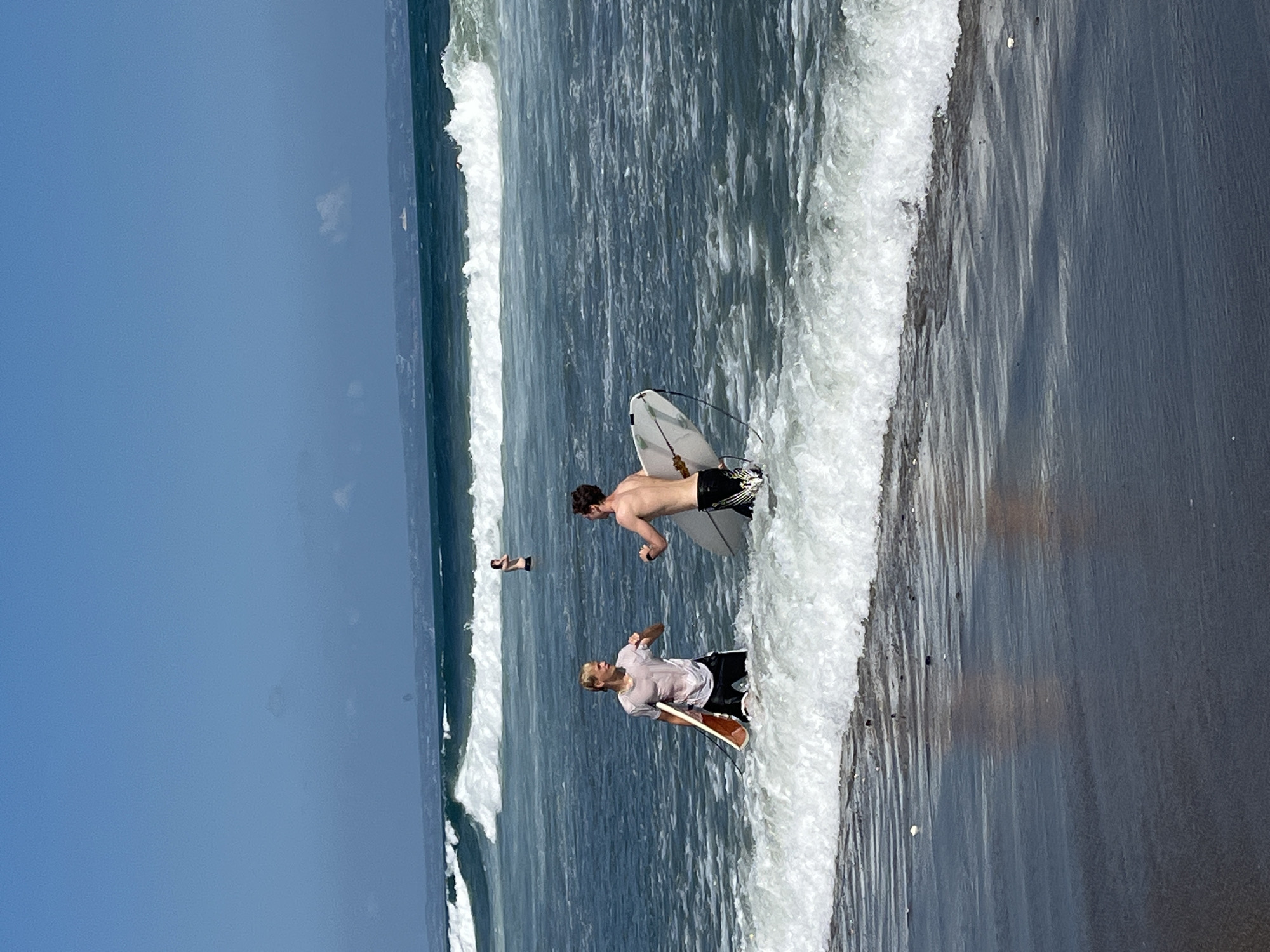 Two young men are in the ocean off the coast of North Kuta, Indonesia, holding surfboards. The man on the left, with blonde hair, is wearing a white t-shirt and black shorts. He holds a light brown surfboard with his right hand. The man on the right, with brown hair, is shirtless and wearing black shorts. He holds a white surfboard with a blue leash. Both men are facing each other and appear to be talking or interacting. In the background, a third person, seemingly a child, stands in the water. The ocean has moderate waves with white foam. The sky is clear and blue. The scene takes place on a beach with dark sand in the foreground.