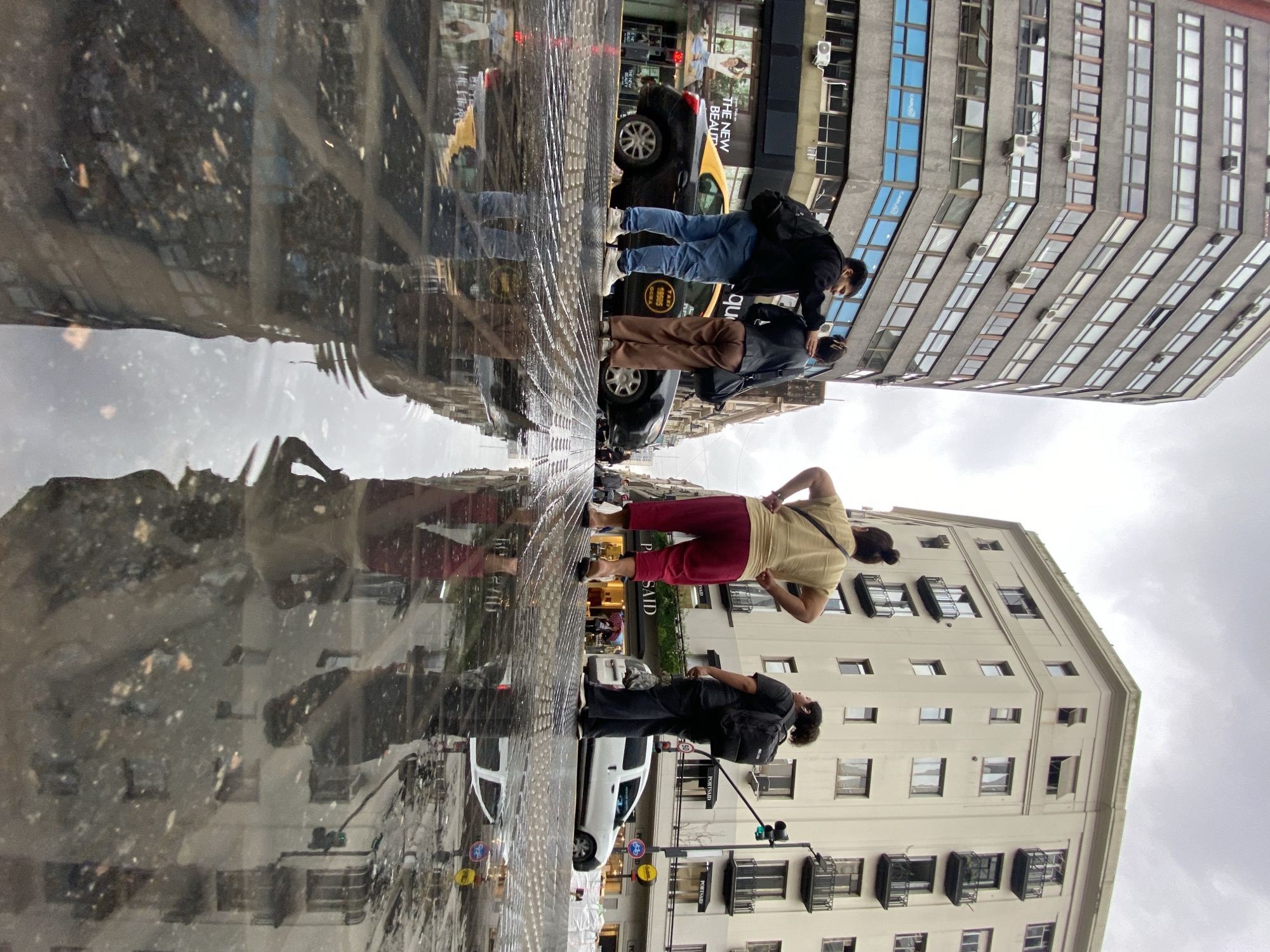 The image depicts a street scene in Buenos Aires, Argentina, reflected in a large puddle. Several pedestrians are standing near parked cars. Two people are on the right, with the closer person wearing a tan shirt and red pants, while the other is wearing black clothing and a backpack. On the left, two other people wearing black jackets and blue jeans stand near a yellow taxi. The background includes buildings with multiple windows. The reflection shows a distorted view of the street and buildings, indicating the presence of water on the pavement. The sky is overcast.