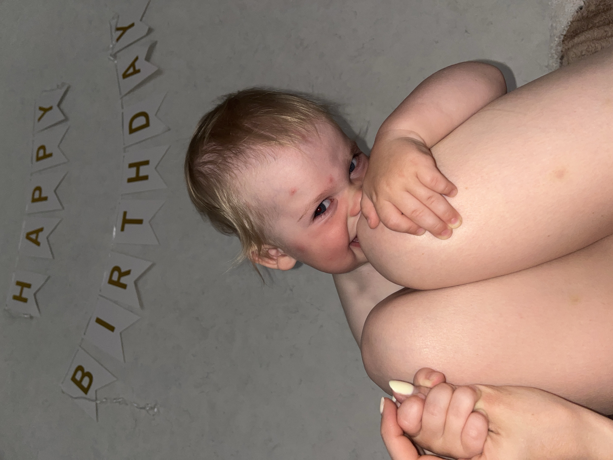 A fair-skinned baby with blonde hair is breastfeeding. The baby has visible red marks on their forehead. A "HAPPY BIRTHDAY" banner is affixed to the wall. A fair-skinned person is also visible. The location is likely in a home, based on the lack of public or outdoor features. It is not possible to confirm that the location is the city of Chaikovsky, Russia.