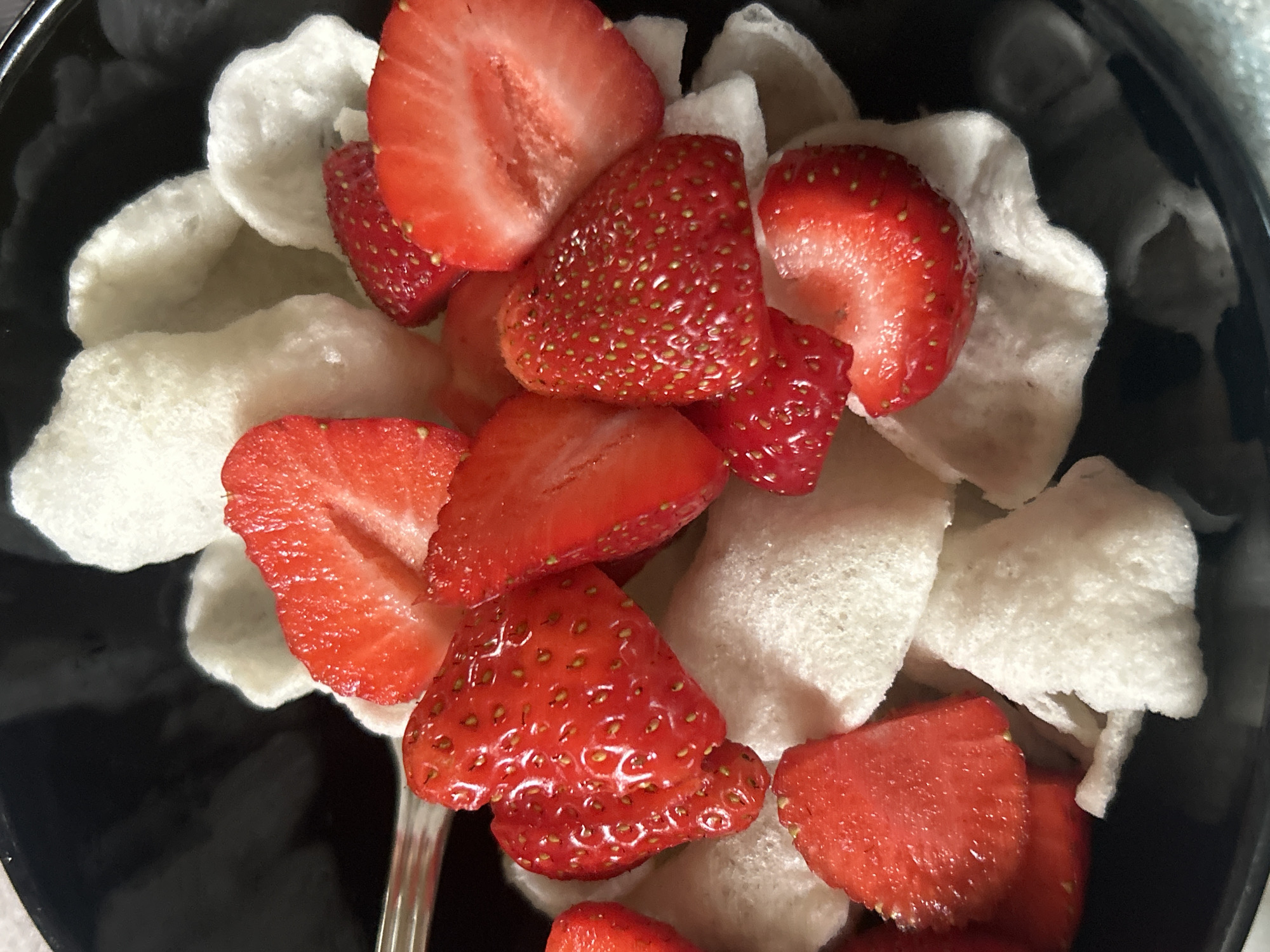 The image shows a black bowl filled with sliced strawberries and white, puffy chips. The strawberries are a vibrant red color and appear to be freshly cut. The chips are irregularly shaped and white, possibly shrimp chips or a similar snack. A silver spoon is visible within the bowl. The location is unknown.