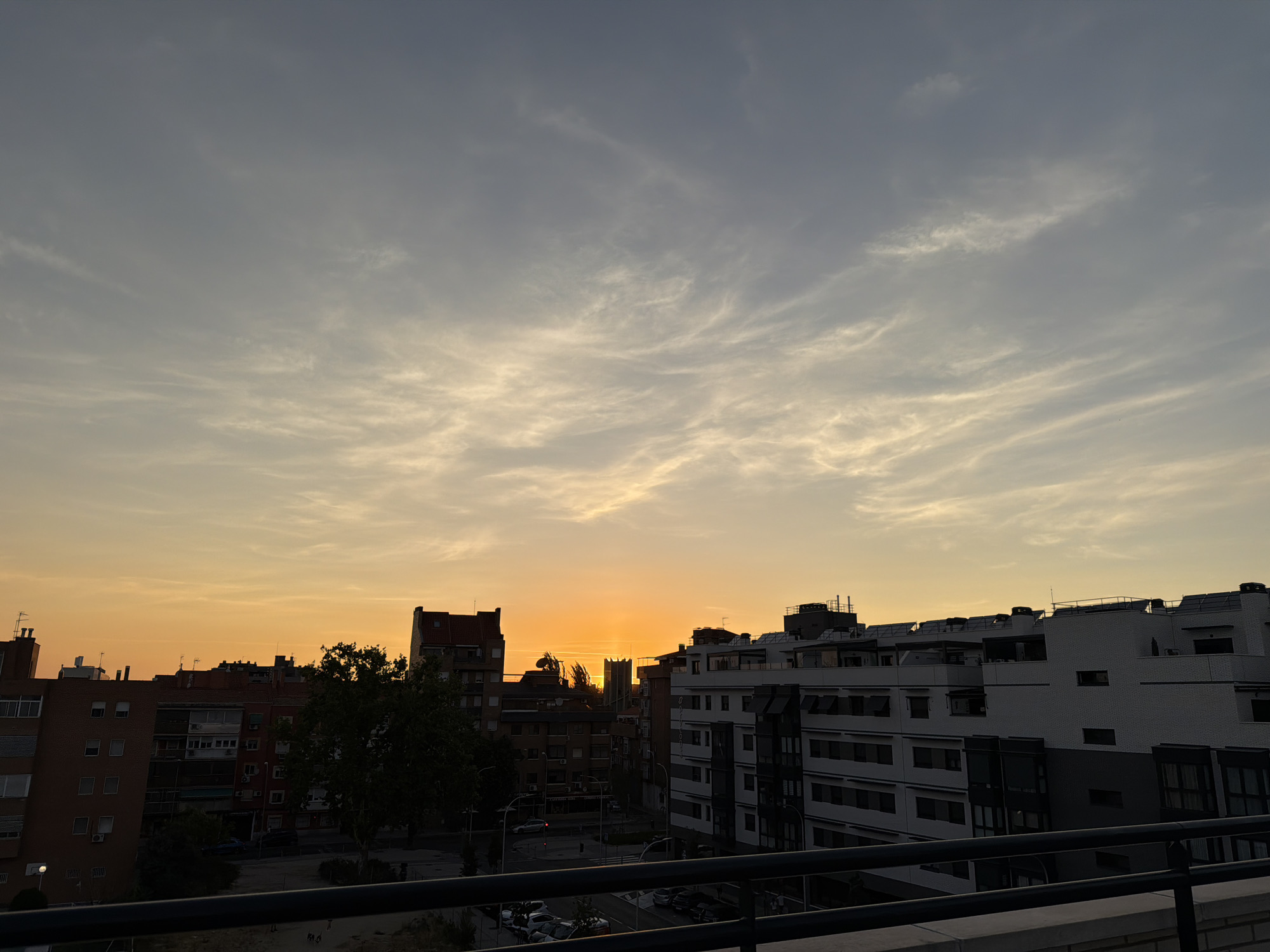 The image depicts a cityscape at sunset, viewed from a balcony. The sky is a gradient of colors, starting with a bright orange near the horizon where the sun is setting and transitioning to a pale yellow and then a light greyish blue towards the top. Wispy clouds are scattered across the sky, catching the sunset light and appearing in shades of white and pale orange.
In the foreground, a dark metal railing of a balcony is visible, suggesting the viewpoint is from an elevated position. Below the railing, there is a blurry glimpse of what appears to be urban ground with some foliage and perhaps the tops of trees.
The middle ground is dominated by a dense collection of buildings. On the left, there are several brick apartment buildings with visible windows, balconies, and rooftop antennas. Some of these buildings are darker in color, while others are lighter. A large, dark green tree partially obscures some of the buildings. Further into the city, a tall, slender structure resembling a church steeple or a tower is visible, silhouetted against the sunset.
On the right side of the image, modern apartment buildings with white and grey facades are prominent. These buildings have multiple floors, visible windows, and rooftop structures. The architectural style appears more contemporary than the brick buildings on the left.
In the background, more buildings stretch into the distance, their shapes softened by the atmospheric haze and the fading light. The overall scene conveys a sense of urban life at dusk, with the warm glow of the sunset casting a peaceful yet dynamic ambiance over the city. No people are clearly discernible in the image.