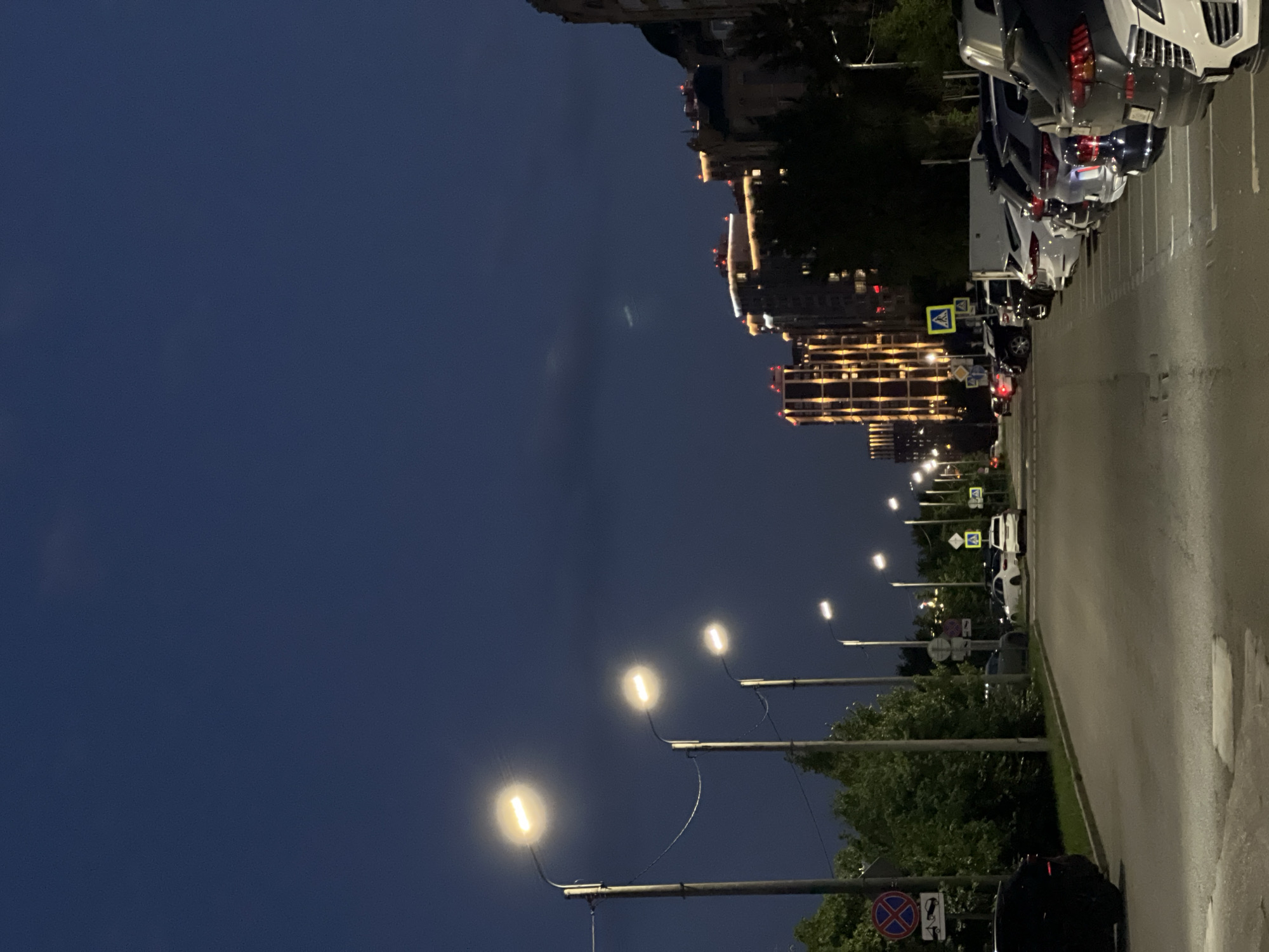 The image shows a street in Kazan, Russia, at dusk. A row of streetlights illuminates the scene, casting a warm glow on the asphalt road and the parked cars. The buildings in the background are modern, with illuminated windows and decorative lights. Trees line the street, their leaves a dark green against the twilight sky. There are no people visible in the image. The overall impression is one of a quiet, urban evening.