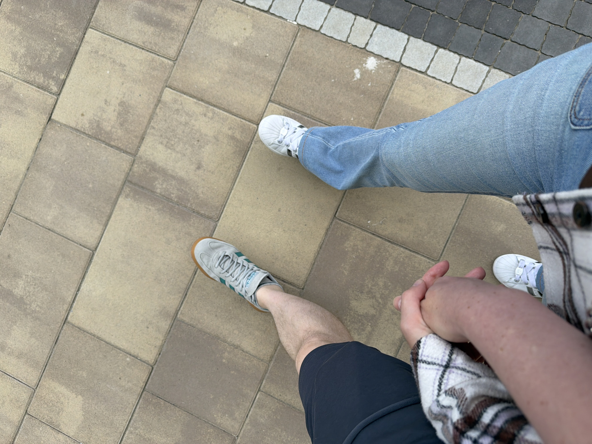 A low-angle shot shows two people holding hands while walking on a paved path. The person on the left wears dark blue shorts and light gray Adidas sneakers. The person on the right wears light blue jeans and white Adidas sneakers. The path is made of rectangular beige and gray paving stones. A border of smaller black and white pavers runs along the right edge of the path. The image is taken from a perspective that suggests the people are walking away from the camera. The environment appears to be an outdoor public space, possibly a park or pedestrian walkway in Kyiv, Ukraine.