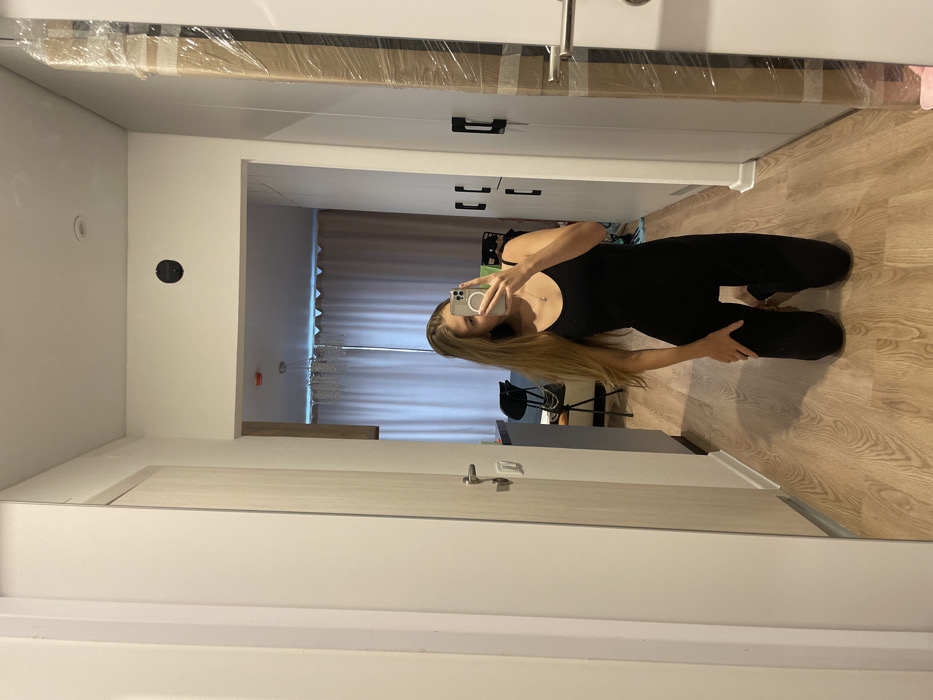 A mirror reflects a woman in a black jumpsuit, kneeling with one hand on her leg and holding a phone up to take a photo. The mirror reflects a hallway with a closed door to the left, and a partially visible room with light-filtering curtains ahead. A black chair can be seen in the room. The floor is light wood-toned. The mirror is framed with white trim at the top and partially at the bottom. A package is wrapped in clear plastic and tape and sits above the mirror.