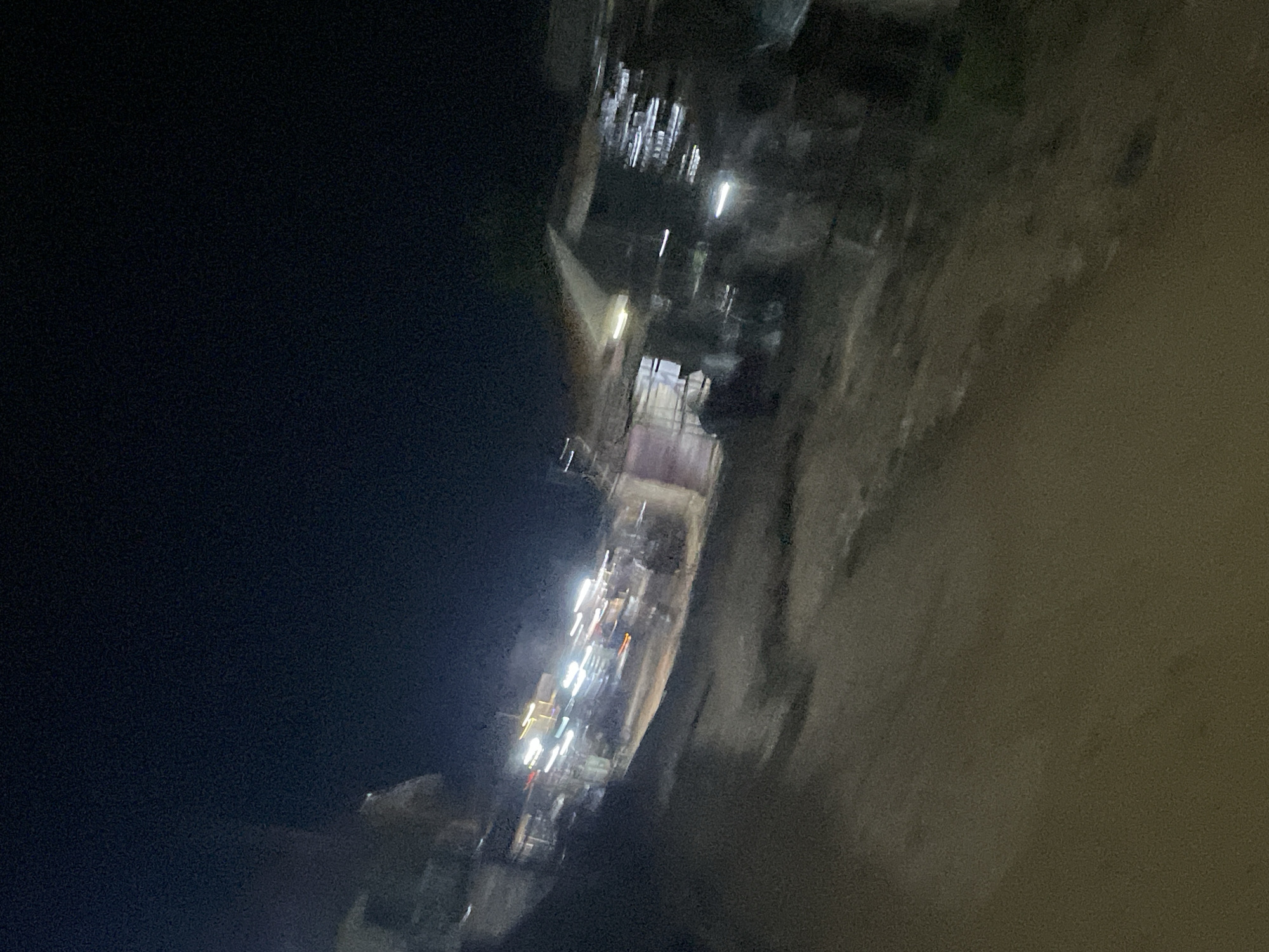 The image is a blurry nighttime street scene, likely in the city of Lokoja, Nigeria. The scene appears to be a marketplace or a commercial area with several structures that resemble small shops or stalls. Bright lights are visible, suggesting illuminated signs or internal lighting from these establishments. The ground is unpaved and appears to be dirt or gravel. The overall impression is of a bustling, albeit somewhat disorganized, nocturnal commercial activity. The blurriness suggests movement, possibly from the camera or passing vehicles. The sky is dark, indicating it is late evening or night.