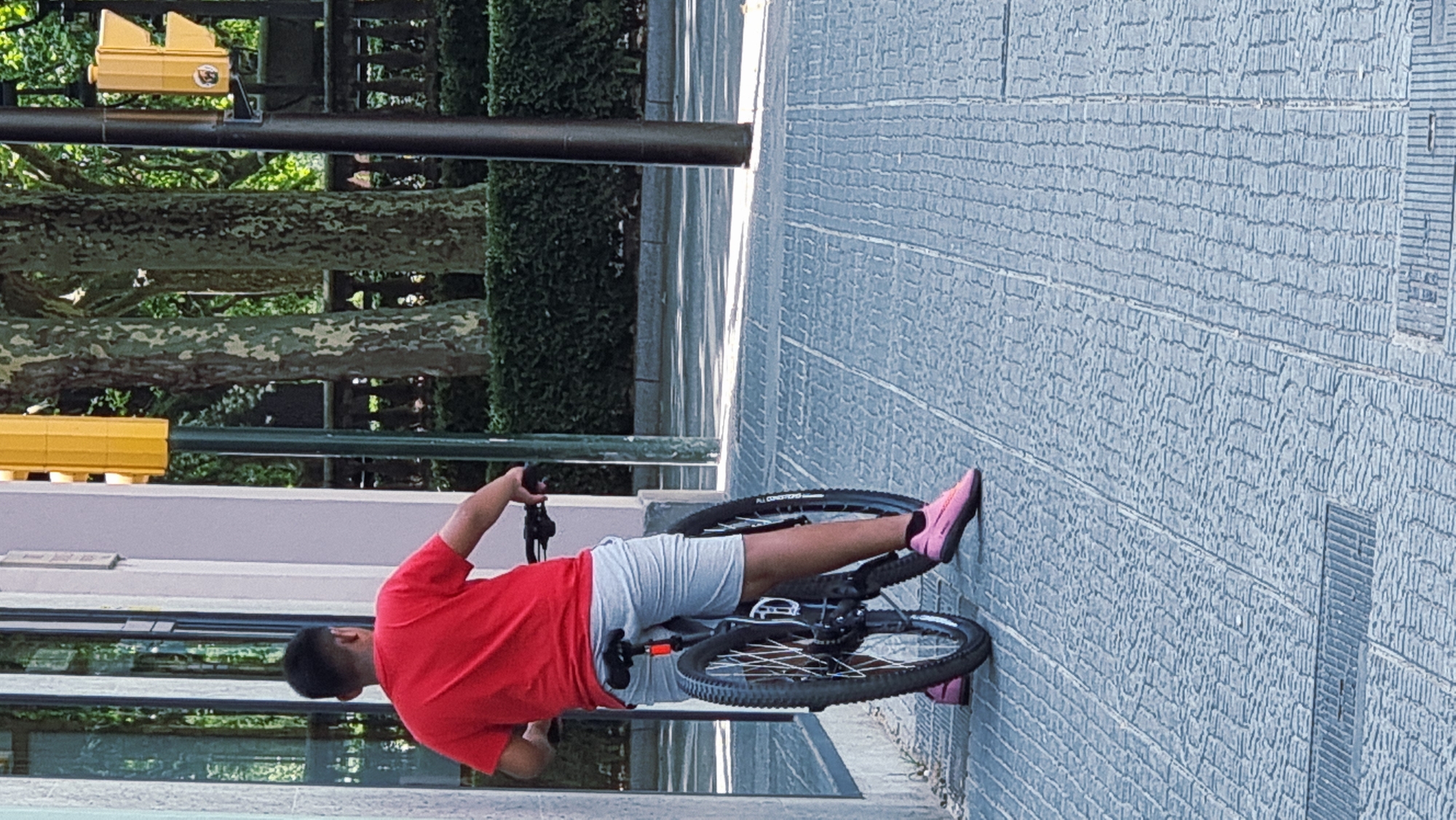 A person is captured in a low-angle shot, seemingly interacting with a bicycle. The individual is wearing a red t-shirt and light-colored shorts, with bright pink slip-on shoes. Their left leg is extended, and their right foot is on the ground, suggesting they might be about to mount or dismount the bike, or perhaps are in the process of performing a stunt. The bicycle itself has a dark frame and knobby tires, indicative of a mountain bike or hybrid.

The setting appears to be an urban environment, characterized by a patterned stone-tiled ground and a textured wall that takes up a significant portion of the background. In the upper left quadrant, there are hints of greenery from trees and bushes, as well as a yellow traffic signal. A dark pole runs vertically through the scene. The lighting suggests it is daytime, likely overcast or shaded, as there are no harsh shadows. The overall atmosphere is casual and possibly recreational. No explicit text is visible in the image.