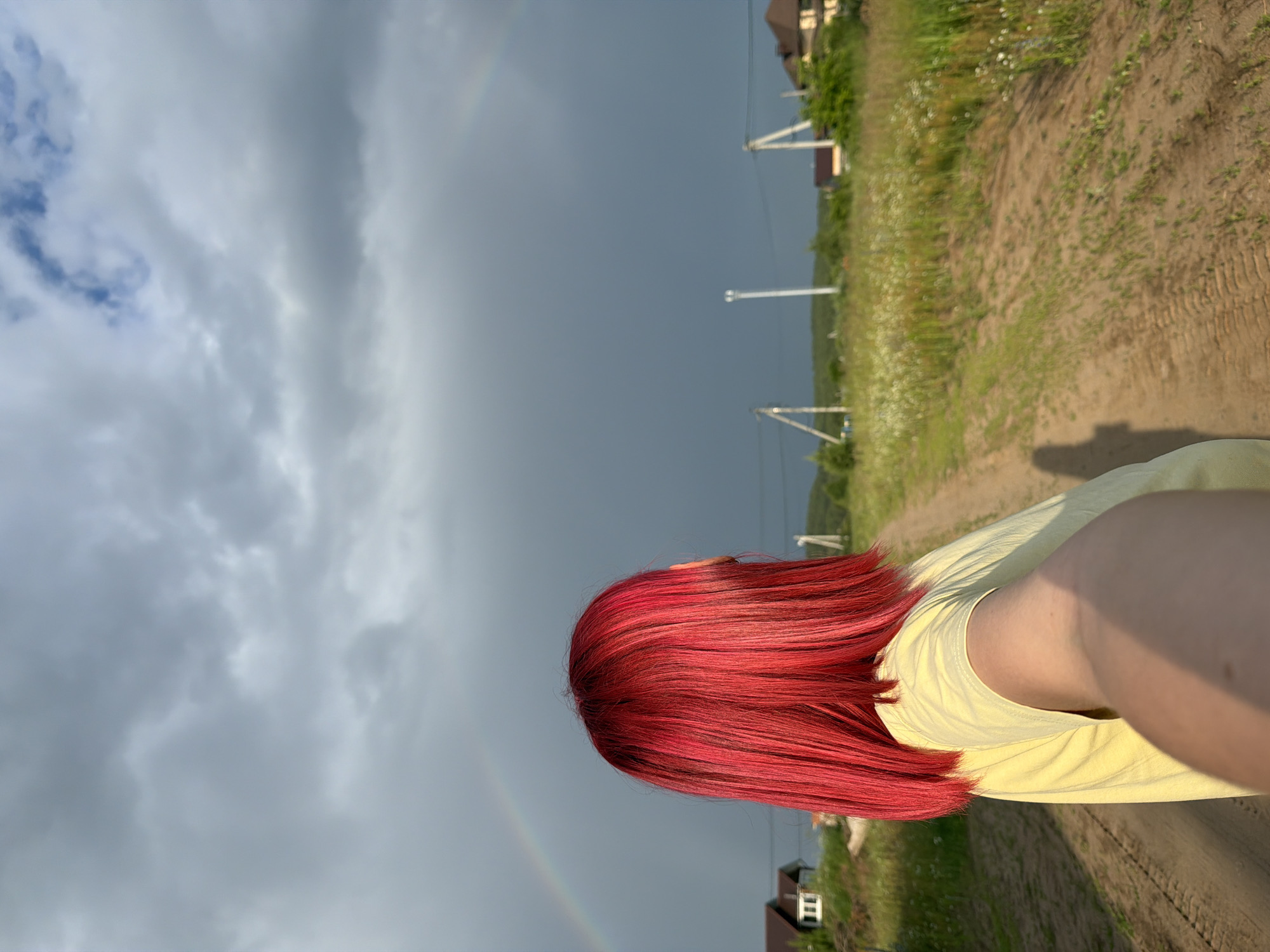 A person with vibrant red hair is facing away from the camera, holding a phone out to take a selfie. They are wearing a light yellow t-shirt. The person is standing on a dirt road in a rural area. In the distance, a faint rainbow is visible against a dark, cloudy sky. Several houses with brown roofs are scattered across the landscape. There are utility poles and wires also visible in the background. The ground is covered with dry grass and some small white flowers. The overall scene suggests an overcast day with a passing shower. The location is likely a dacha settlement or a rural outskirts area, possibly within the city of Chaikovsky, Russia, given the context.