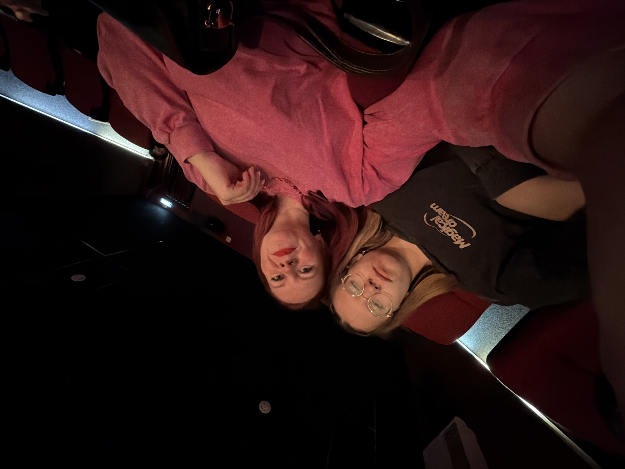 Two women are posing for a selfie in a darkened theater. The woman on the left has red hair and is wearing a pink shirt and red lipstick. The woman on the right has blonde hair, glasses, and is wearing a black t-shirt with "Magical dream" written on it. The theater chairs are red. The location is unknown.