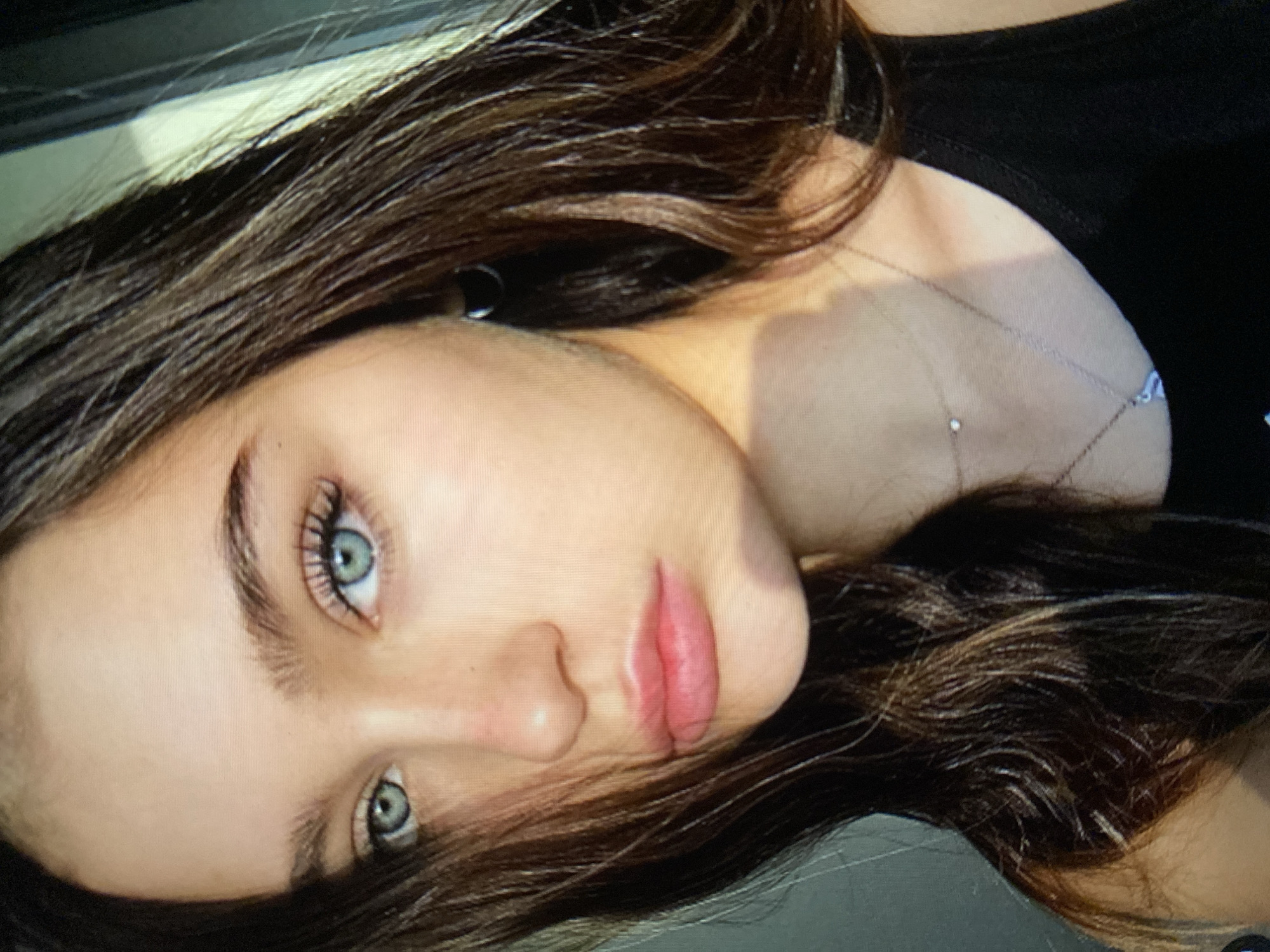 A close-up, head-and-shoulders portrait of a young woman with brown hair and striking blue eyes is captured in natural light. She is looking directly at the camera, her expression neutral. Her hair is dark brown and slightly wavy, framing her face and falling over her shoulder. She is wearing a black tank top and a delicate necklace with a small pendant. The background is softly blurred, with hints of a window or door on the right side, suggesting an indoor setting. The lighting creates a subtle shadow across her right cheek and neck.