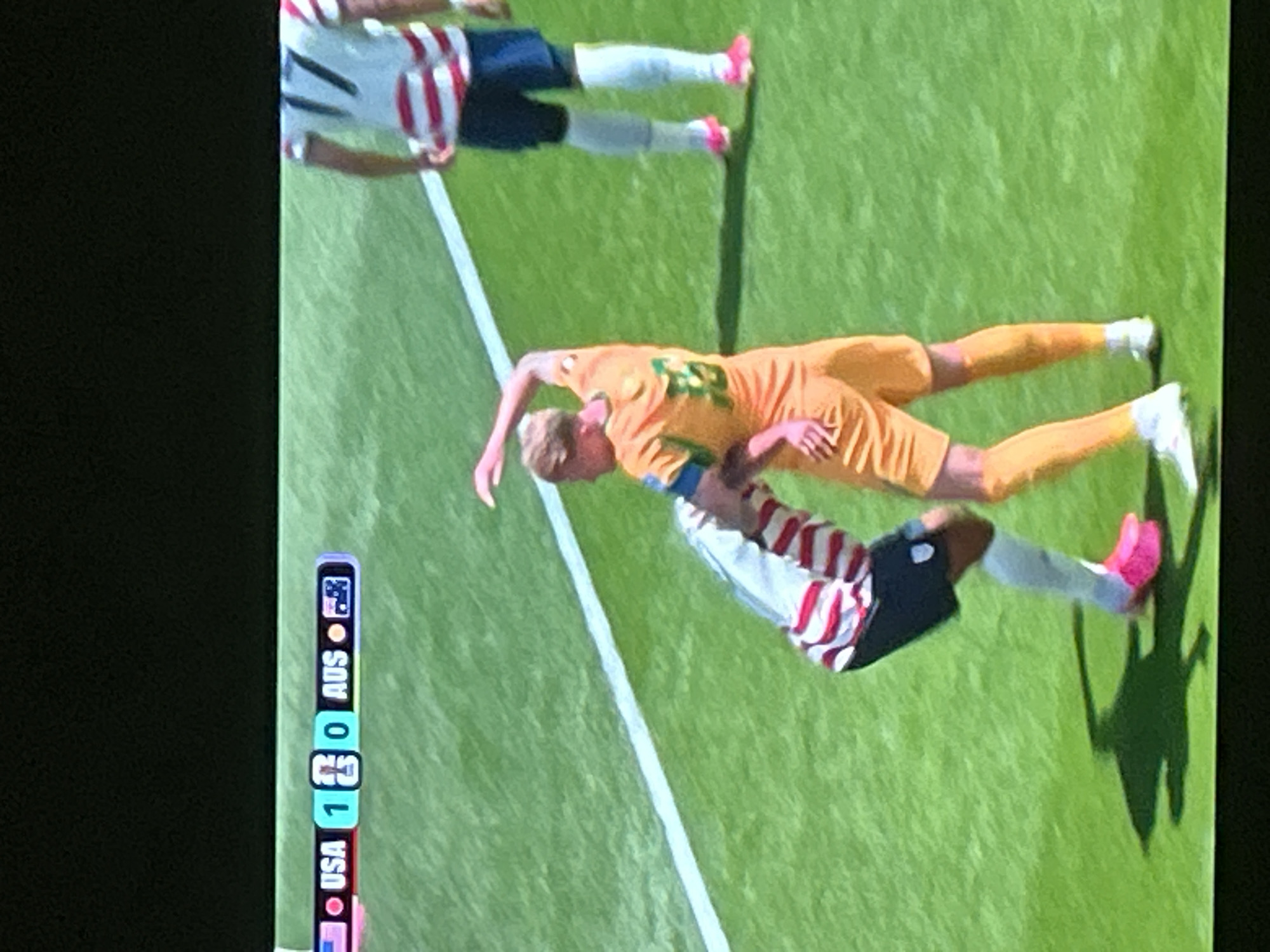 The image shows a close-up view of a soccer match on a green field. A scoreboard in the upper left corner displays "USA 1 0 AUS". Two players are in the foreground, one wearing a white jersey with red and blue stripes and black shorts, the other in an orange jersey and shorts. The player in the orange jersey is reaching out to the player in the striped jersey, who is bent over. Another player in a striped jersey with the number 17 is visible in the background. The lighting suggests it is daytime, with shadows cast on the field.