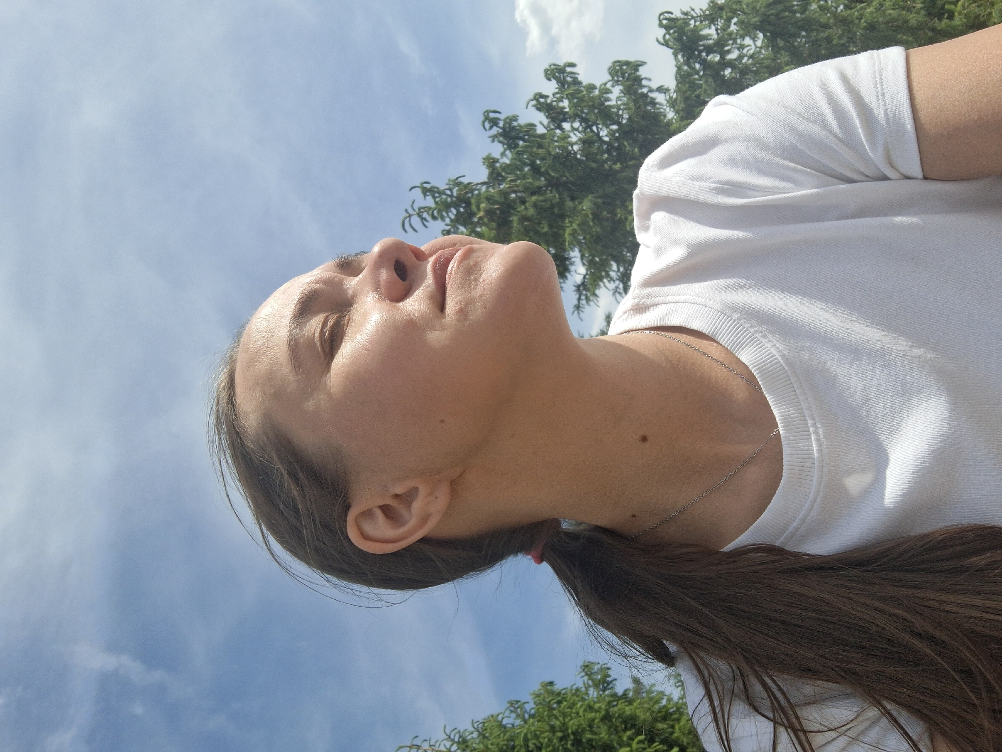 A person is looking up at a sky with clouds and trees in Ольховка, Russia. The person is wearing a white t-shirt and a necklace. Their hair is pulled back in a ponytail with a pink hair tie. The sky is blue with some white clouds. There are green trees in the background.