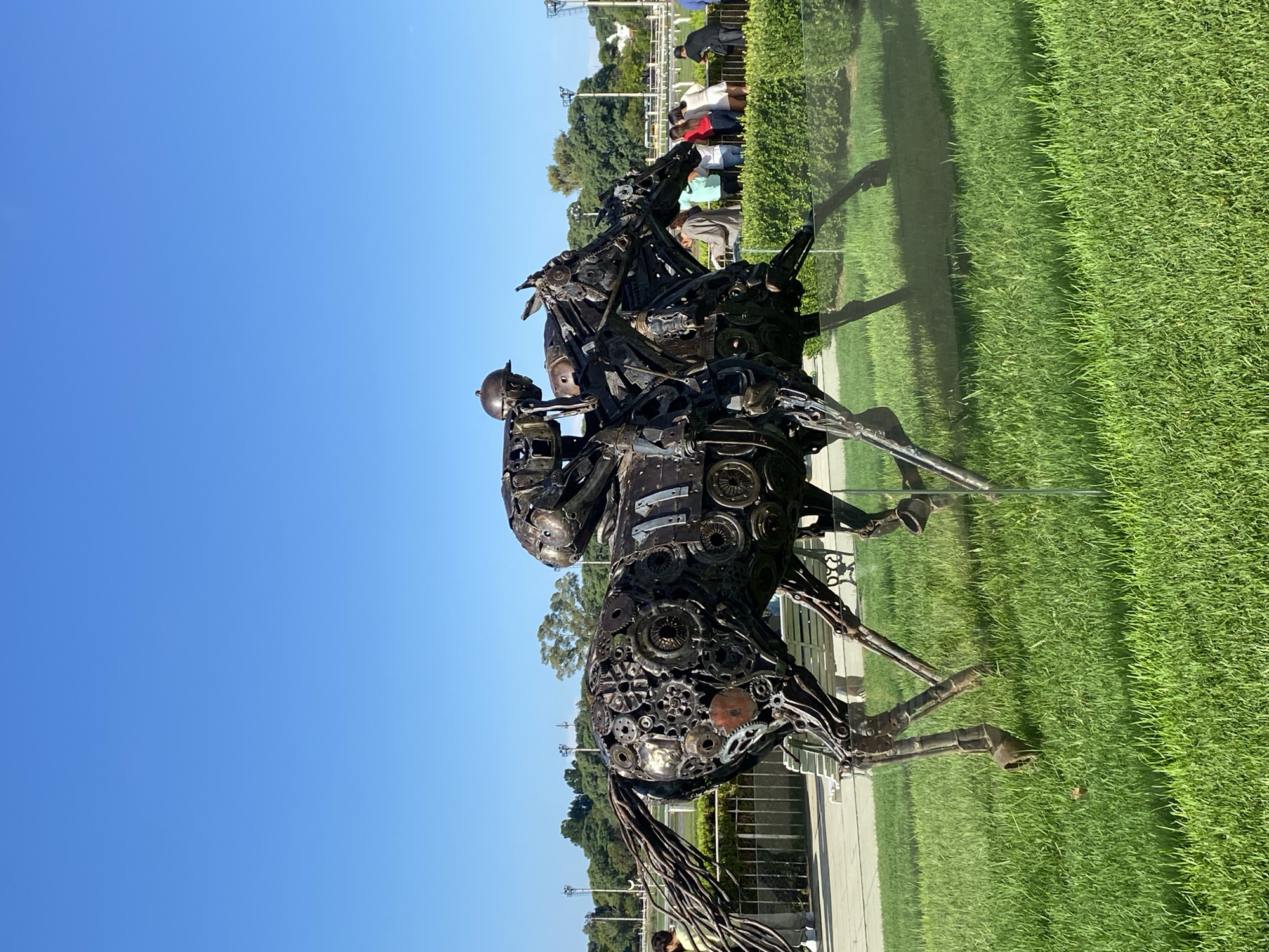 The image shows a metal sculpture of a jockey on a horse, located outdoors in Buenos Aires, Argentina. The sculpture is constructed from various mechanical parts, including gears and pipes. It stands on a patch of green grass against a backdrop of a clear blue sky. In the background, a group of people can be seen standing behind a barrier, with trees and buildings visible further in the distance. The image appears to be taken at eye level, giving a clear view of the sculpture and its surroundings. The lighting suggests it is daytime.