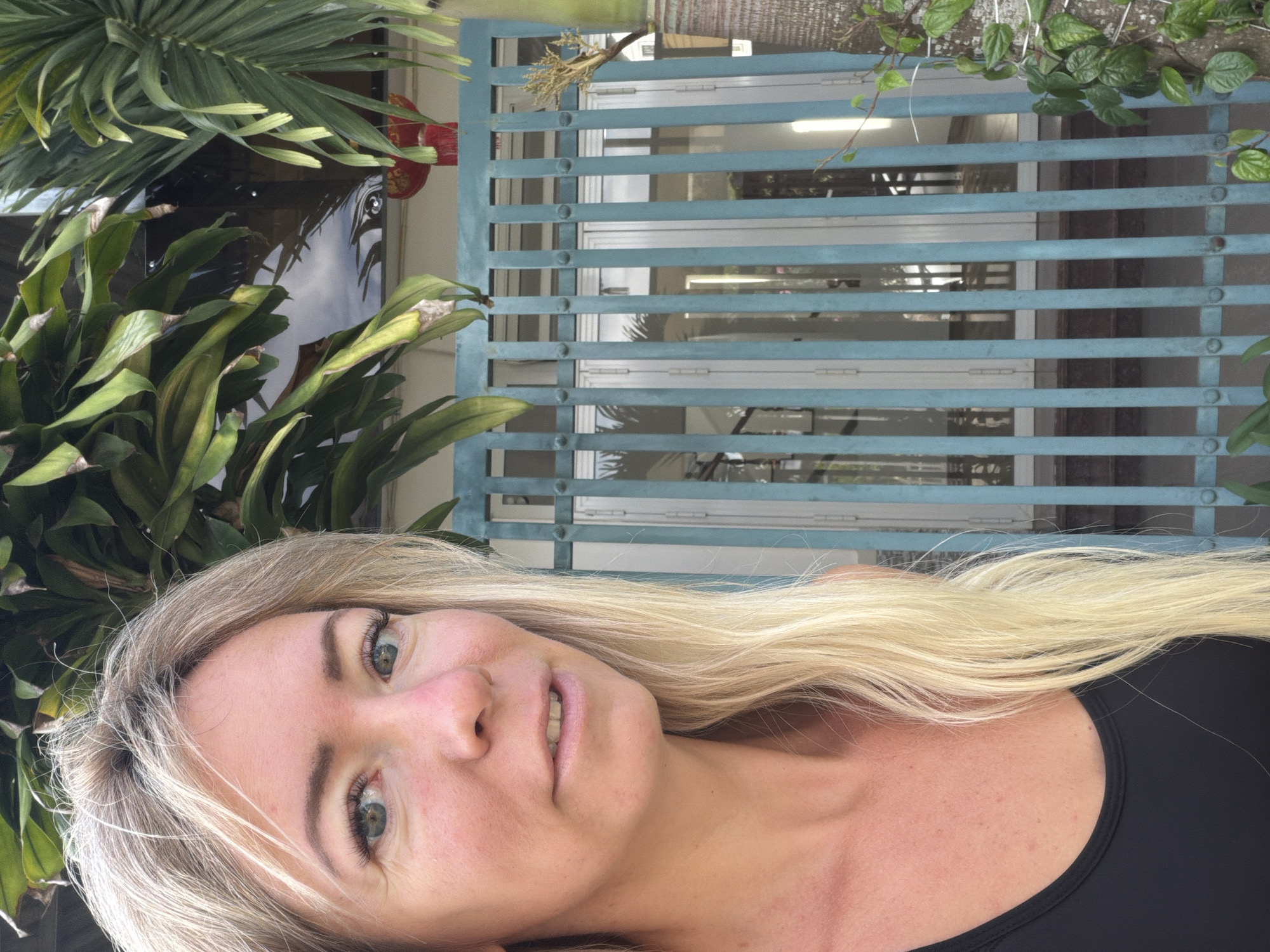 A medium shot captures a woman with long blonde hair against a backdrop of lush greenery and a teal metal gate with vertical slats. She is wearing a black sleeveless top, and her face is turned towards the camera, with her eyes looking slightly to the left. The background features a building with glass doors behind the gate, and a red lantern hanging from an overhang. The scene is set outdoors, likely in a residential area or a place with significant vegetation. The lighting suggests it is daytime, with sunlight filtering through the leaves. The presence of tropical plants indicates a warm climate, consistent with Da Lat, Vietnam.