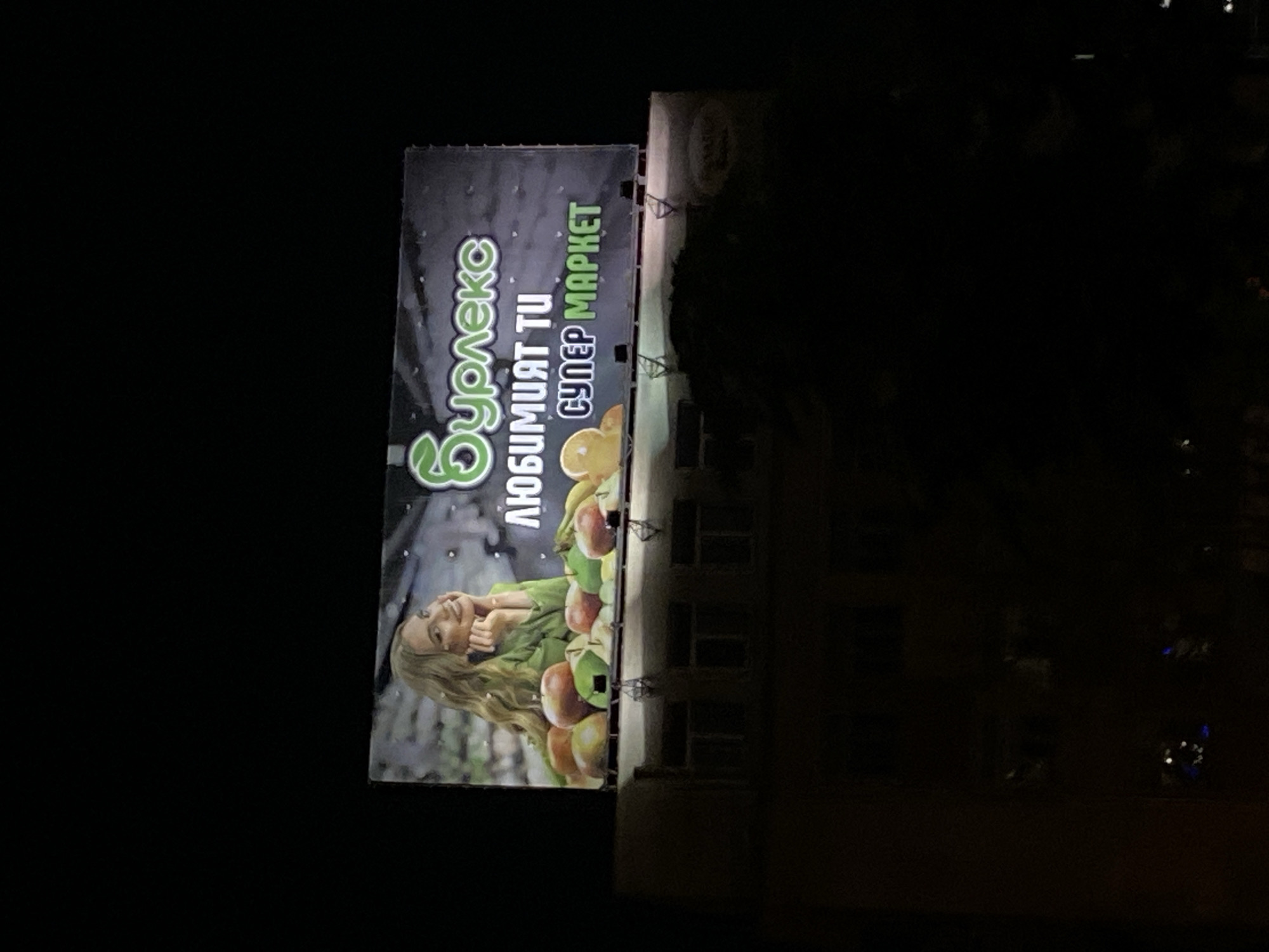 A vertically-oriented photograph shows a billboard in Varna, Bulgaria, situated on the side of a building at night. The billboard promotes the supermarket "Burlesto," displaying its name and slogan in Bulgarian. A woman is featured alongside fruits, possibly apples. The surrounding environment is dark, with only the lit billboard and faint building details visible. The building has multiple windows and a flat roof. The photograph is taken from a distance, offering a medium shot of the billboard in relation to the building.
