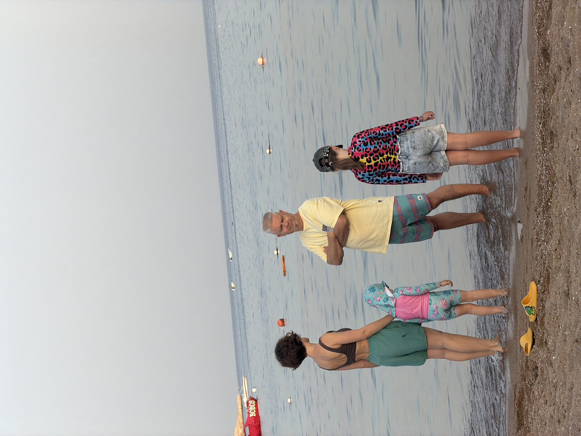 This is a vertical, full-shot photograph of four people on a beach, with the water of the Red Sea in the background. The scene appears to be set in Sharm El Sheikh, Egypt, given the location context.
The subjects are a man in a yellow t-shirt and patterned shorts, a young girl in a leopard-print top and denim shorts, a woman in a dark top and green shorts, and a small child in a patterned rashguard and pink life vest. The man is standing with his arms crossed, looking towards the water. The young girl is also looking out at the water, with a baseball cap on. The woman is facing away from the camera, and the small child is partially obscured behind her, facing the water.
There are several buoys or markers visible in the water. On the beach to the right, there is a pair of yellow sandals. In the lower left corner, a red object with white text is partially visible, appearing to be part of a rescue float. The sky is overcast, suggesting a potentially cooler time of day or simply cloudy weather. The lighting is soft, and the water is calm. The overall mood seems relaxed, possibly indicating a family outing or a break from activities. The text "BOD" and "SOS" is visible on the red object.