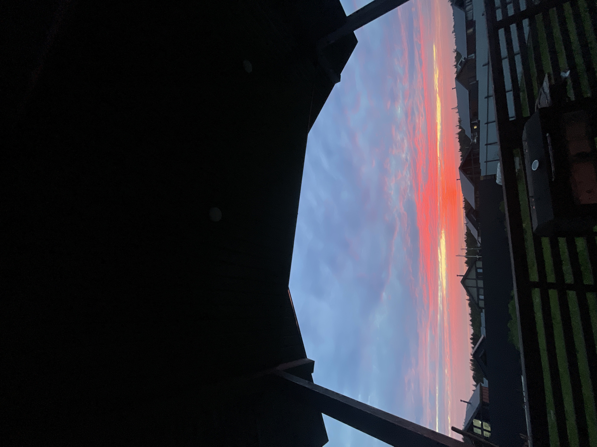 The image depicts a sunset viewed from a covered porch or balcony, looking out over a village or suburban area in Podshivalovo, Russia. The sky is filled with vibrant colors of pink, orange, and blue, with wispy clouds stretching across the horizon. In the foreground, a dark, angular structure, likely the roof and support beams of the porch, frames the view. Below the sky, a row of houses with dark roofs and some with illuminated windows are visible. A dark wooden fence runs across the midground, and beyond it, green grass and the silhouettes of trees create a natural backdrop. A black barbecue grill is partially visible on the porch in the lower right corner. The overall scene suggests a peaceful evening in a rural or semi-rural setting.