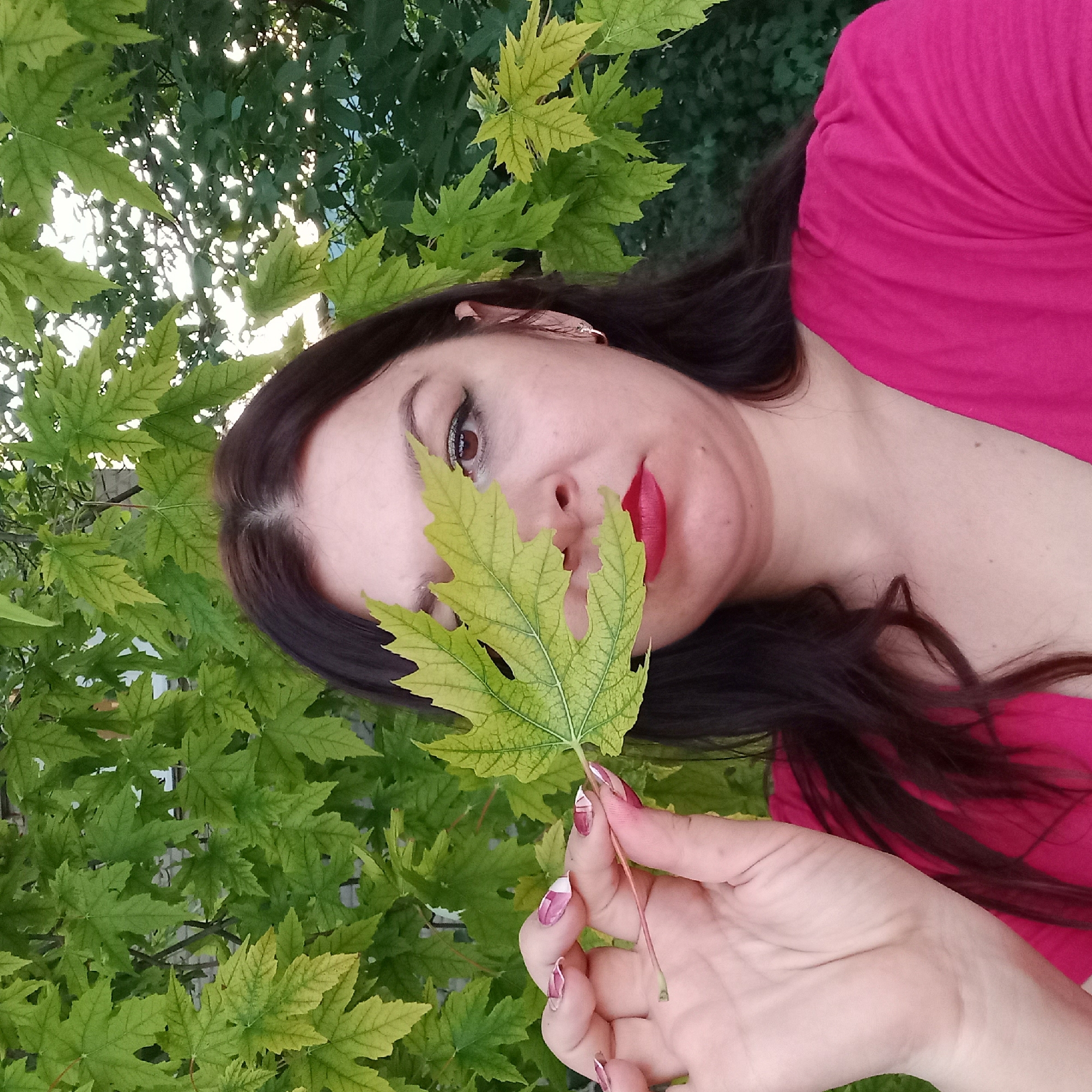 A woman with long, dark hair, wearing a bright pink top, is holding a green leaf in front of her face, obscuring her left eye and part of her nose. Her right eye is visible, with makeup including eyeliner and eyeshadow. She has bright red lipstick on. The background consists of green leaves, some of which are tinged with yellow, suggesting an outdoor setting in the city of Berezhynka, Ukraine. The lighting appears to be natural daylight.