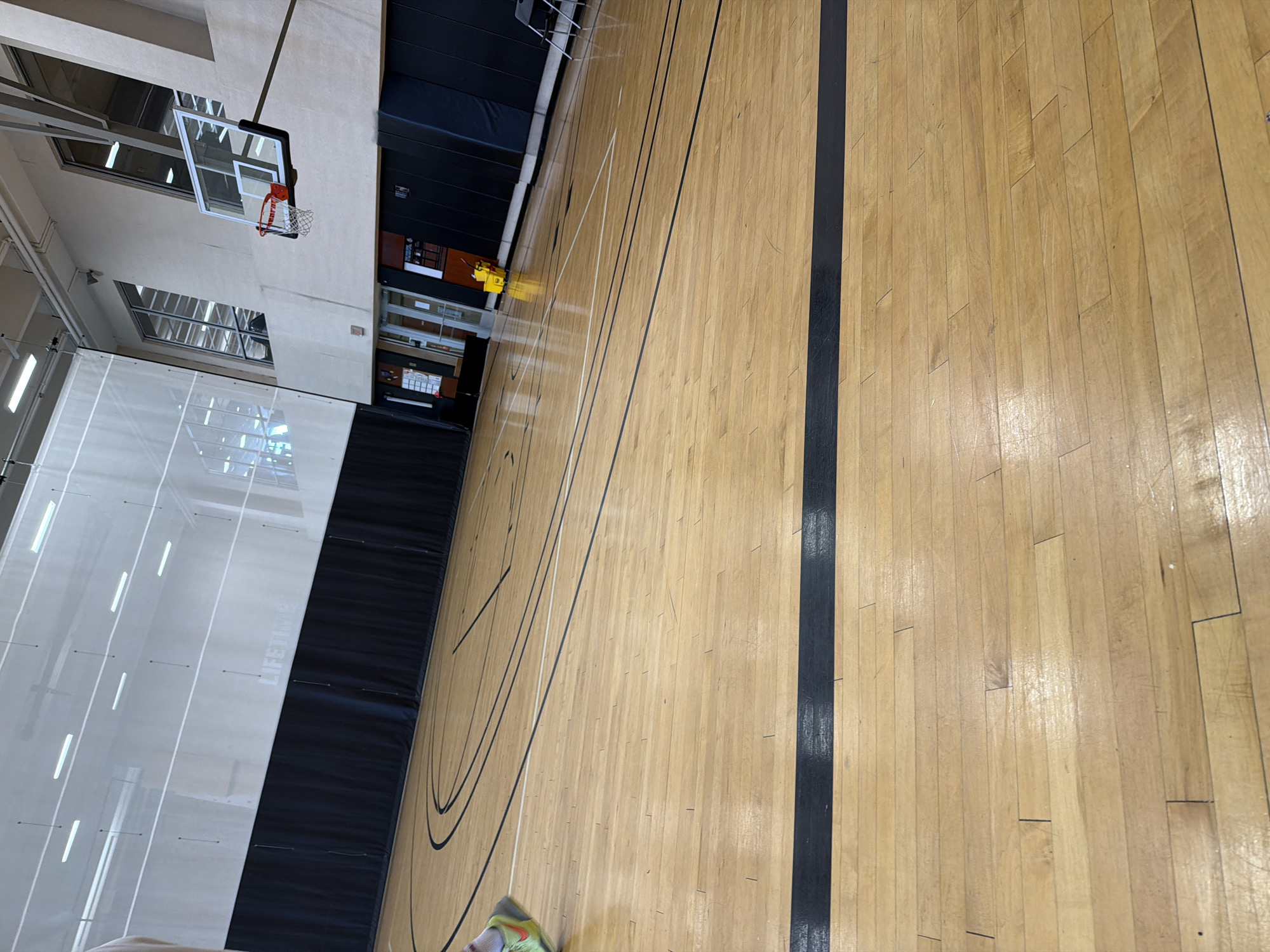 This image captures an indoor basketball court at a Life Time Fitness center, located in Beachwood, Ohio, United States of America. The court features a polished wooden floor with distinct black and white lines marking various play areas. A basketball hoop, equipped with a clear rectangular backboard and a red rim, is positioned on the right side of the frame.
Along the far wall, black padded panels line the lower section, above which a white translucent fabric or mesh barrier extends towards the ceiling. The "LIFETIME" logo is faintly visible on this white barrier, roughly centered in the upper-mid section of the background. Multiple horizontal fluorescent or LED light fixtures are visible through the translucent material overhead. On the left side of the image, the lower leg and a bright neon yellow-green athletic shoe of an individual are partially visible, cropped by the frame.
Towards the right side, beyond the basketball hoop, the wall features large, multi-paned windows angled upwards. Below these windows, an entryway with double doors is visible, and a bright yellow mop bucket with a wringer stands nearby. Further right, against another section of the black padded wall, two grey folding chairs are present. The court is well-lit, with the lighting creating reflections on the wooden surface.
