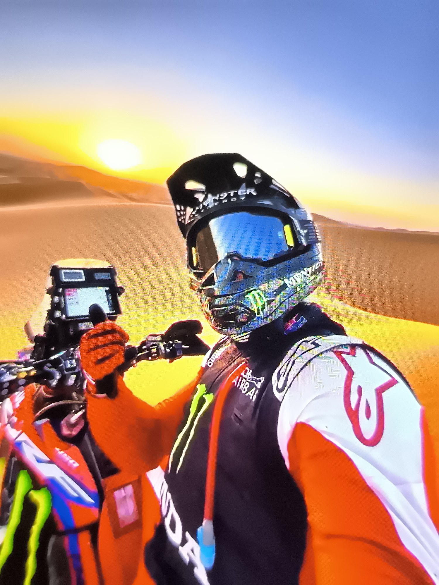 The image shows a person wearing a black and blue helmet with Monster Energy logos and blue goggles, and an orange and white racing suit with Monster Energy branding. They are holding onto the handlebars of a dirt bike in a desert landscape with sand dunes. The sun is low in the sky, casting a warm glow over the scene. The motorcycle has a navigation device mounted on the handlebars. The image does not provide any information about the city of Bălți, Moldova.