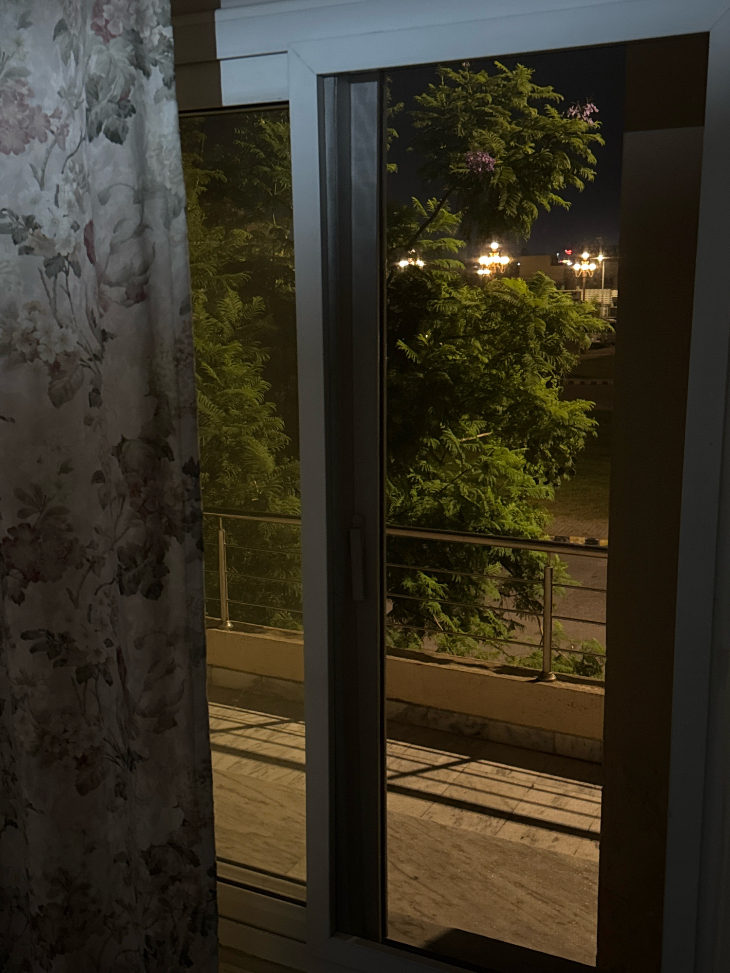 The image shows a view through a window at night, with a curtain on the left side. Outside, there are trees with green foliage and some purple flowers. Several streetlights are visible, illuminating the scene. A balcony with a metal railing is also present. The ground outside appears to be paved, with shadows cast by the railing. The overall environment suggests an outdoor space within a residential area, possibly in Kohistan Enclave, Pakistan, given the context. There are no people visible in the image.