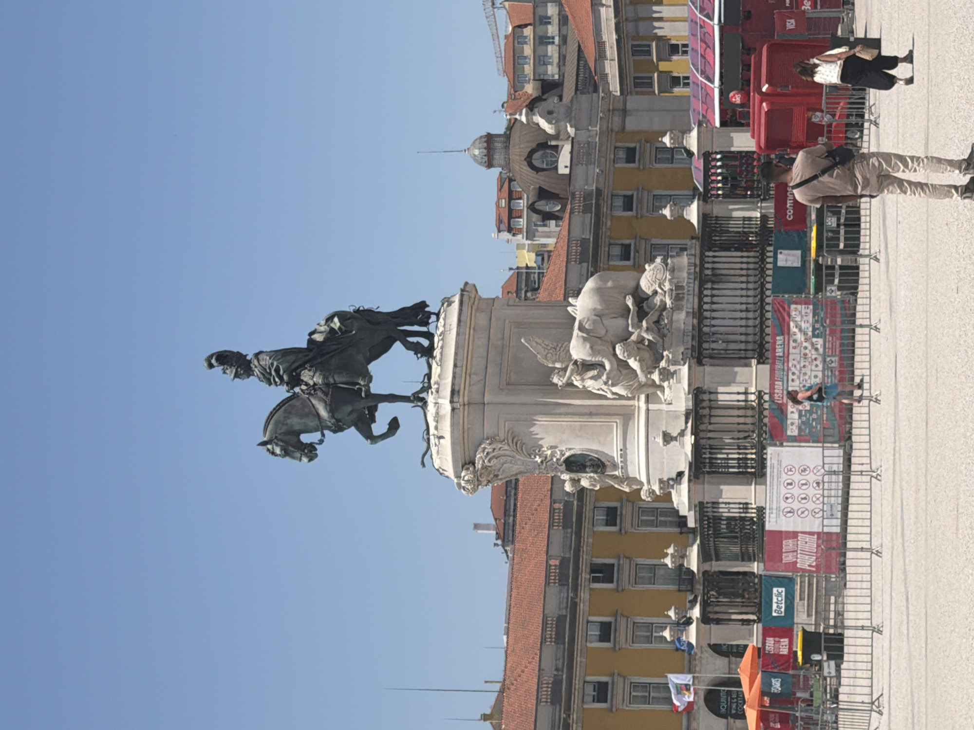 The image shows a sunny day in Lisbon, Portugal, featuring the equestrian statue of King Dom Jose I in the center of Commerce Square. The statue stands atop a large white marble pedestal adorned with sculptural reliefs. The background reveals yellow-colored buildings with red tile roofs, characteristic of Lisbon's architecture. In the foreground, several people are present, including a man in light-colored clothing facing away from the camera, and a woman in a black skirt and white top also facing away. Fencing and banners, some advertising "LISBOA FOOTBALL ARENA" and "Betclic", are visible, suggesting an event or public gathering. A red tour bus is also partially visible on the right side of the frame. The sky is clear and blue.