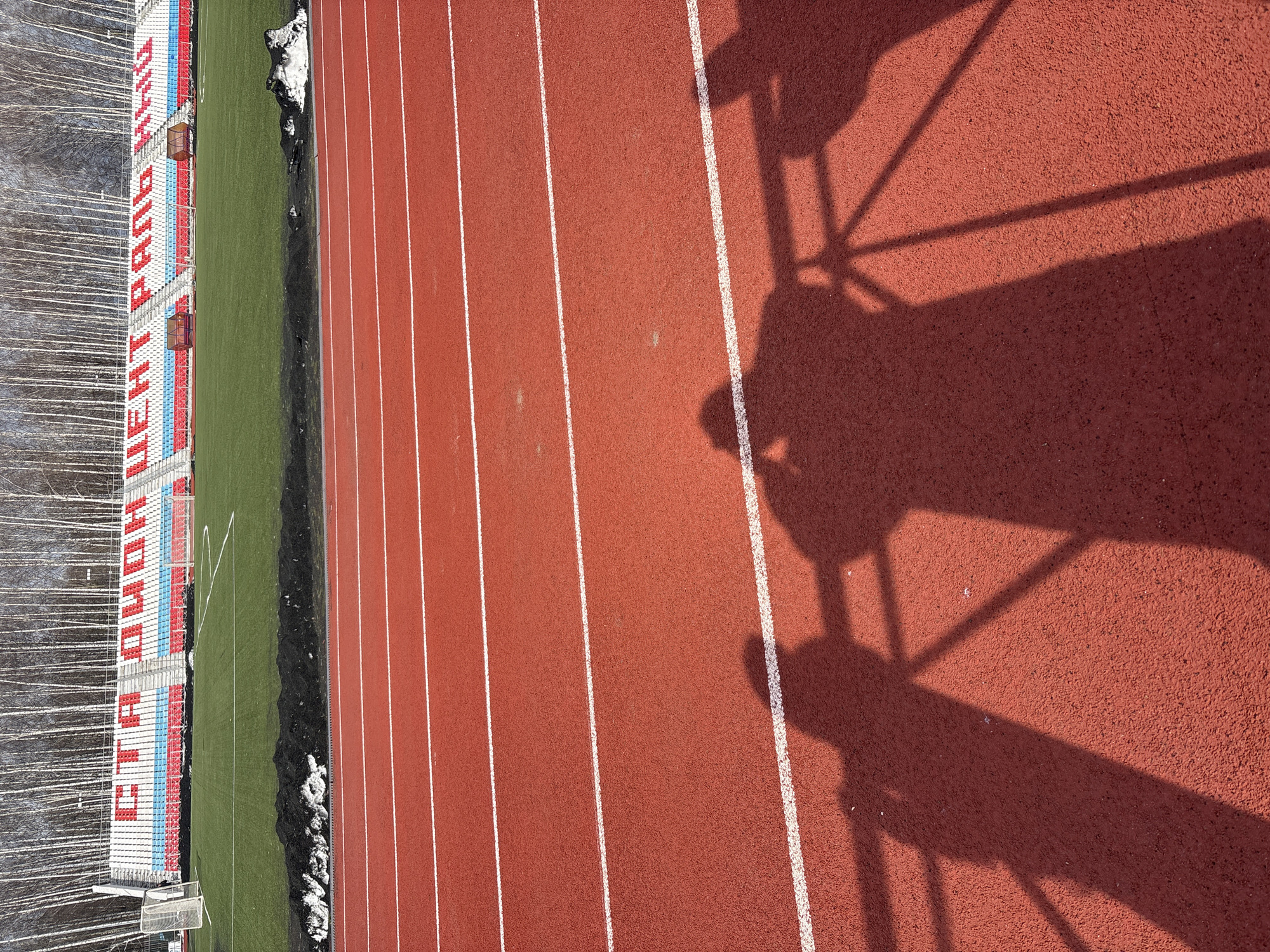 The image depicts an outdoor athletic track adjacent to a football field and stadium seating. The track, rendered in a reddish-brown color, is divided into lanes by white lines. The seating area features rows of blue, red, and white seats. Snow is visible near the edge of the track and football field. Shadows are cast on the track surface, suggesting a specific time of day. The lettering on the stadium identifies the location as the "Центральный стадион" (Central Stadium), located in the city of Chaikovsky, Perm Krai, Russia.