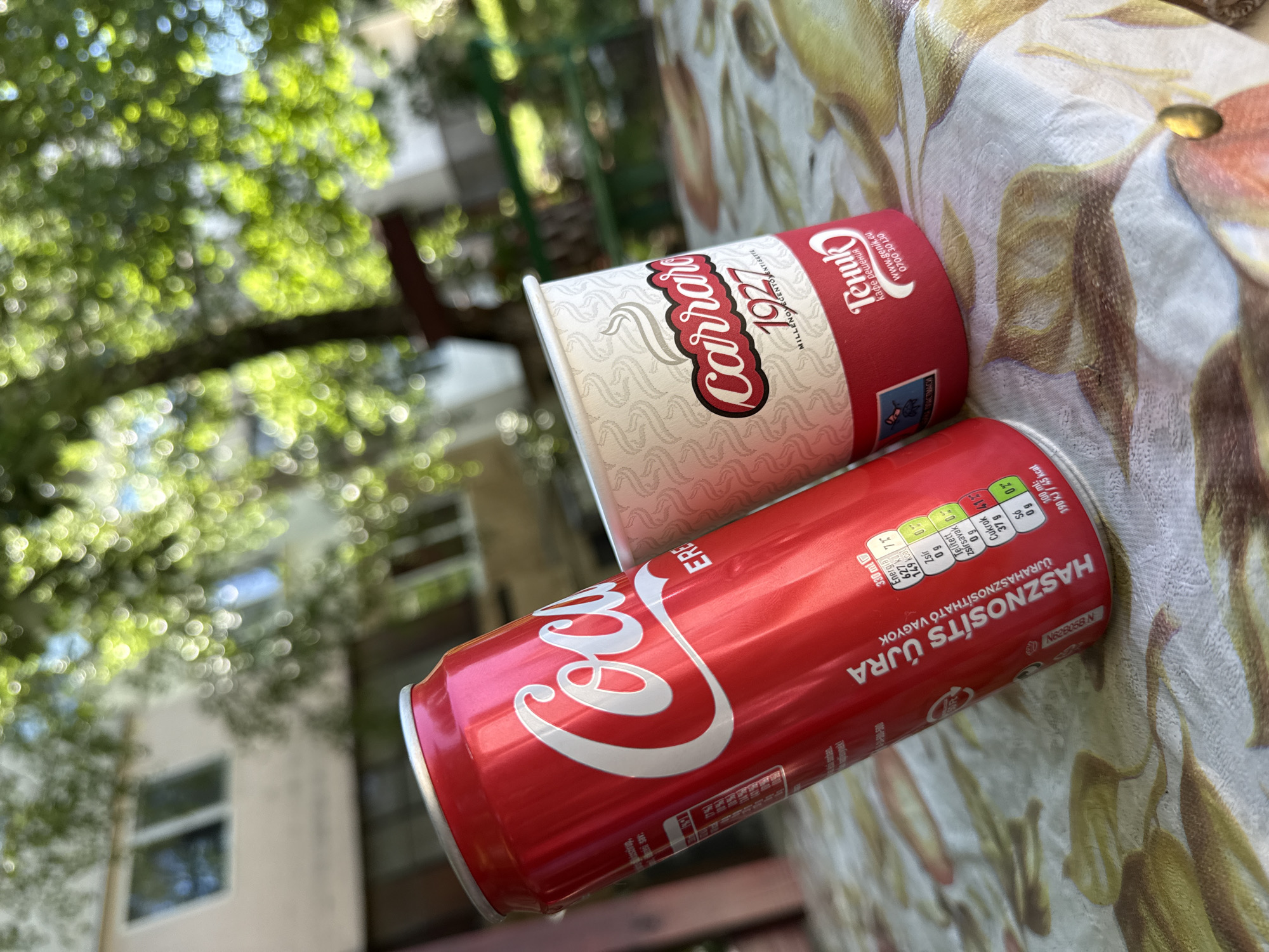 Two beverage containers, a red Coca-Cola can and a white and red paper cup with the Carraro 1927 logo, are placed on a table covered with a floral-patterned tablecloth. The Coca-Cola can is tilted, showing the brand's iconic logo and nutritional information in Hungarian. The paper cup displays the Carraro logo and the text "MILLENOVECENTOINTESSENSTE." Below the Carraro logo, the cup also features the "Тепик" logo and website "WWW.TENIK.EU". The background is softly blurred, revealing green foliage, a building with windows, and sunlight filtering through the leaves, suggesting an outdoor setting. The scene is likely captured in Vratsa, Bulgaria, given the presence of the Russian text on the cup.