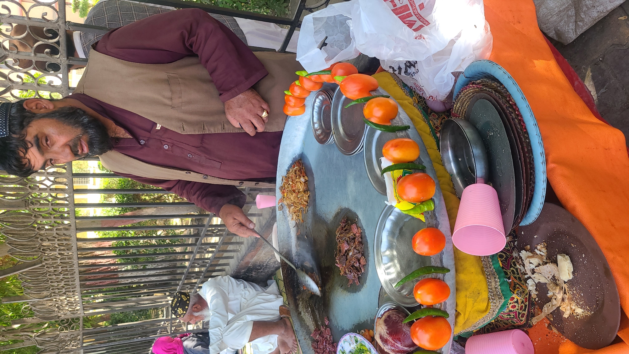 A street vendor in Quetta City Tehsil, Pakistan, prepares food. A man wearing a maroon shirt and a tan vest stirs food on a large, round griddle. Tomatoes and green chilies decorate the edge of the griddle. Plates and pink cups are stacked nearby. Another man sits on the ground. A fence and building are visible in the background.
