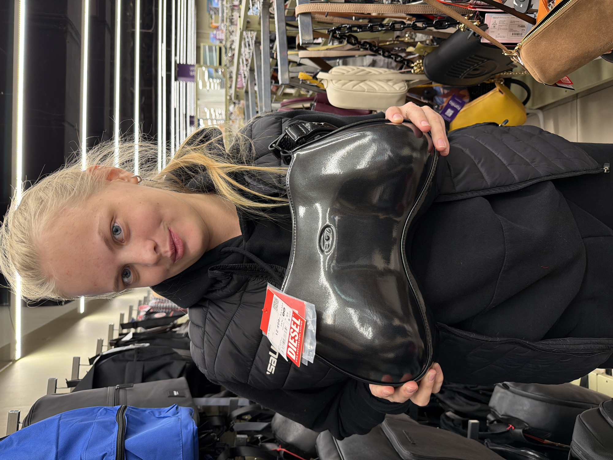A young, light-skinned woman with blonde hair is holding a shiny, dark gray purse in a retail setting. She is wearing a black hooded jacket and looking toward the camera. The purse has a red tag attached, which reads "DIESEL". Behind the woman are rows of bags on shelves. Above, there are other purses and accessories hanging. The location is likely a store, as indicated by the merchandise on display. The store is in Izhevsk, Russia, according to the tag on the bag.