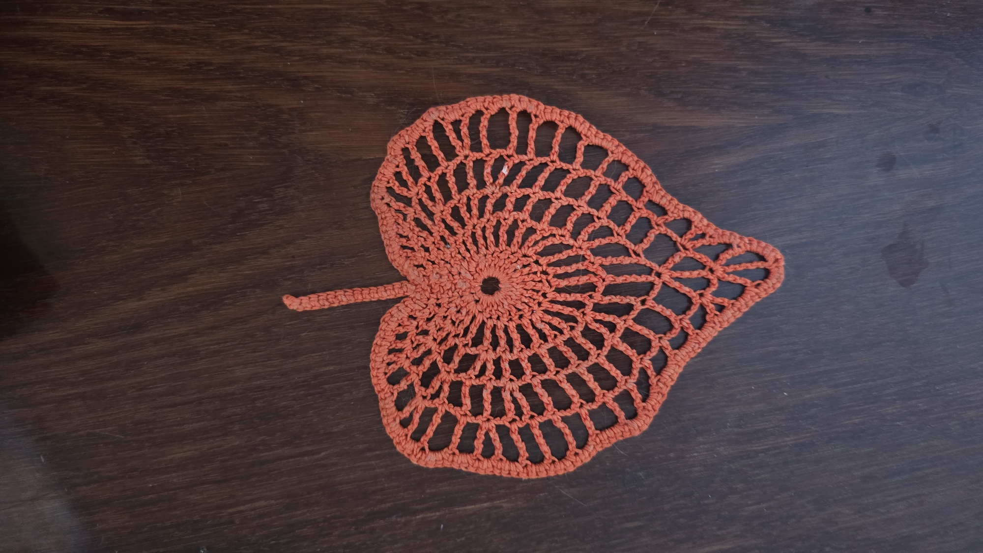 An orange crochet doily shaped like a heart with a stem lies on a dark wooden table. The doily has an open, web-like pattern radiating from a central hole. The table surface has a visible wood grain and a few dark spots. The image was taken in Novohuivynske, Ukraine.
